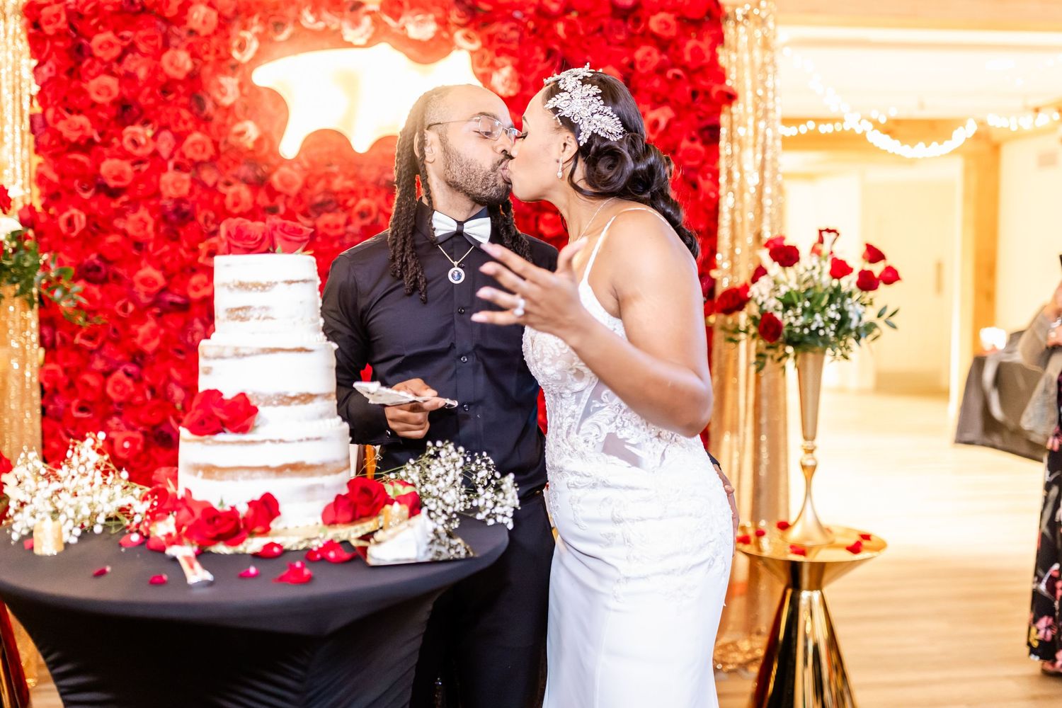 Wedding couple sharing a playful cake-cutting moment in front of a vibrant red flower wall.