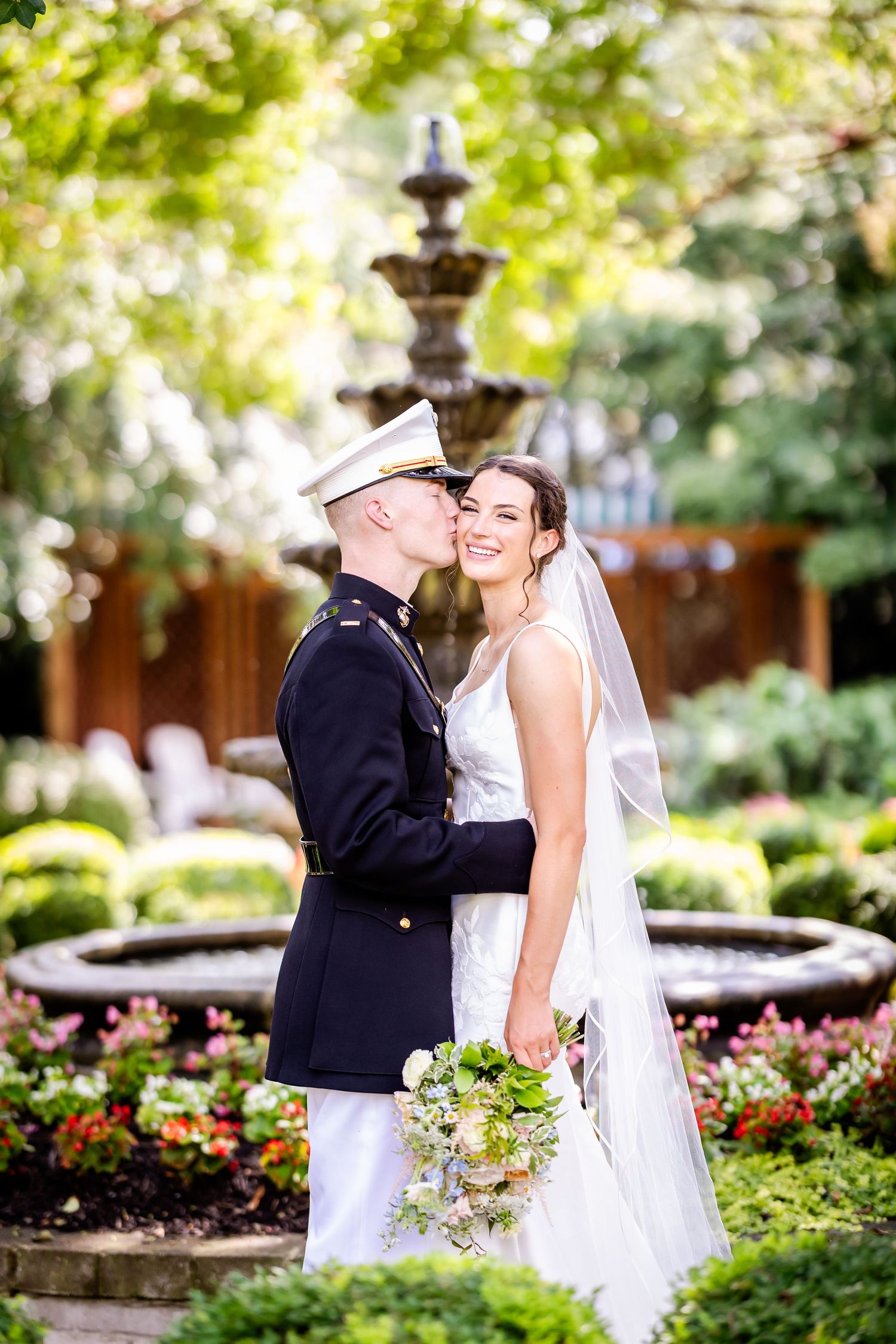 Military wedding couple shares romantic moments in a lush garden with fountains.