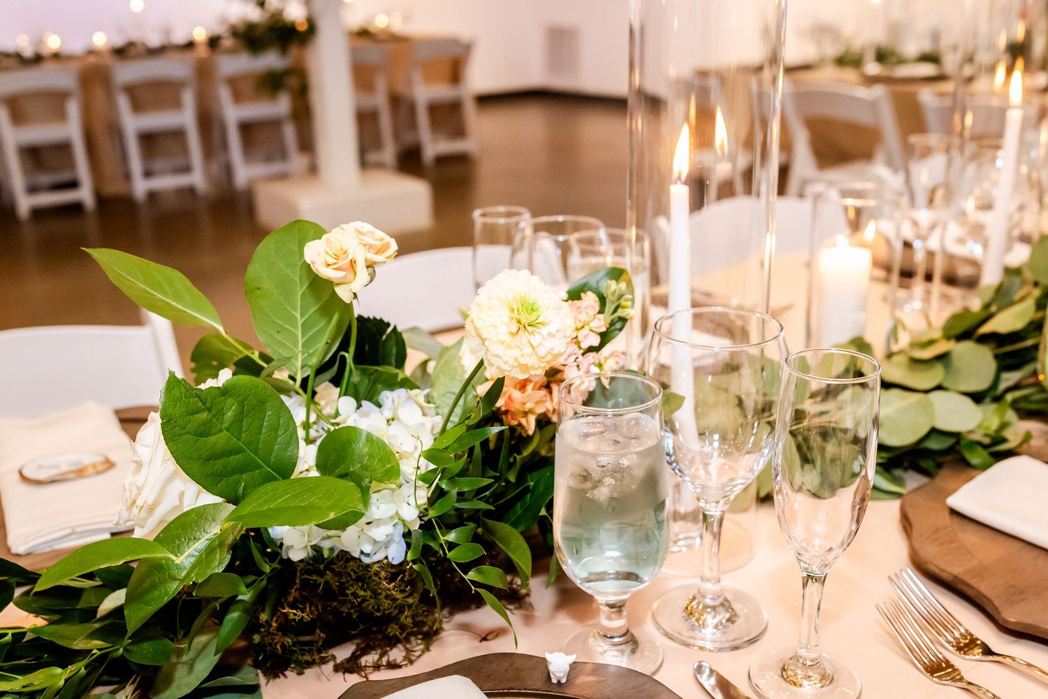 Elegant wedding reception table with lush greenery, white and peach floral centerpiece, candles, and glassware on a neutral tablecloth.