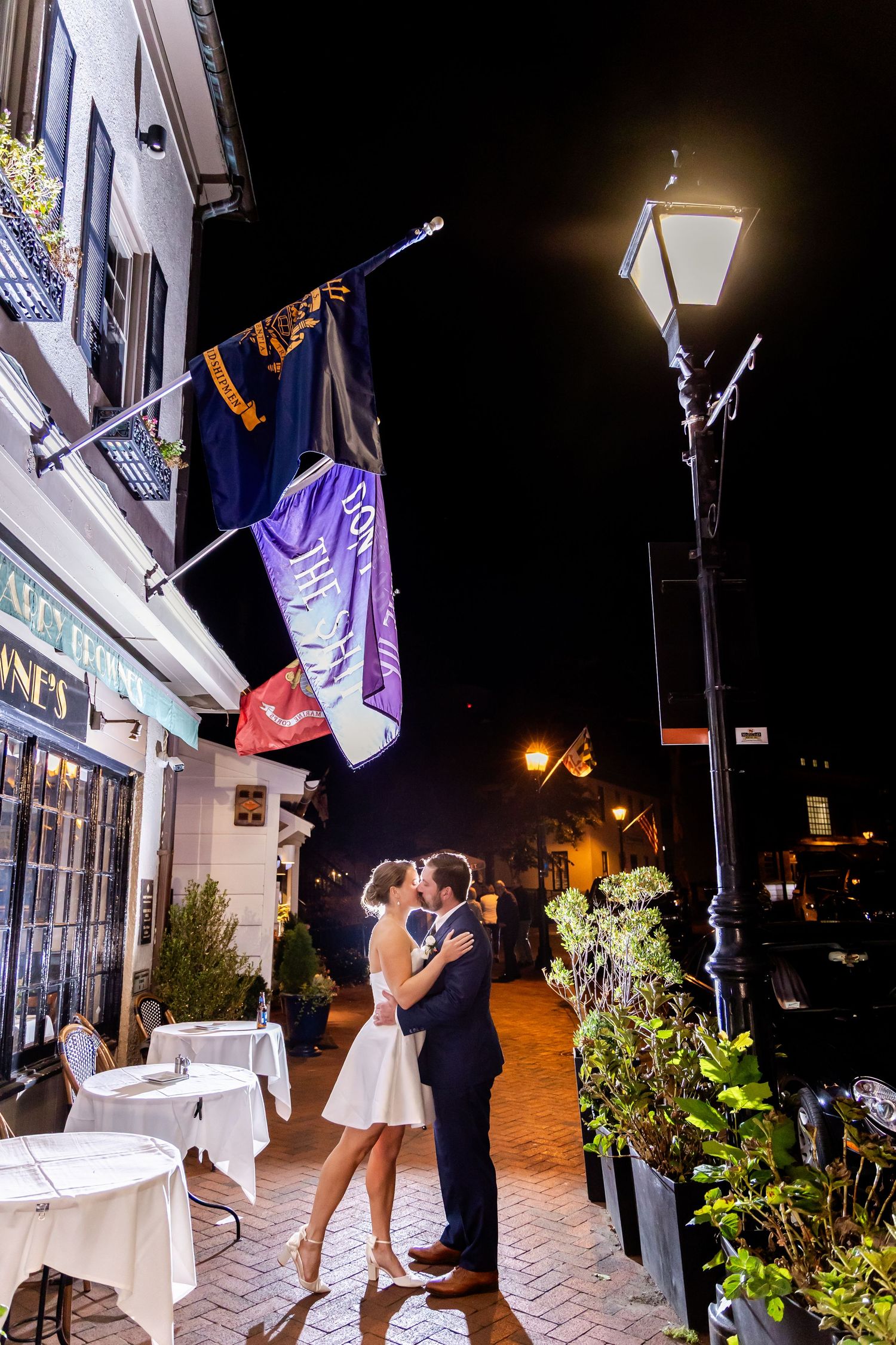 Couple sharing a romantic kiss on a cobblestone street at night, surrounded by flags and a glowing lamppost.