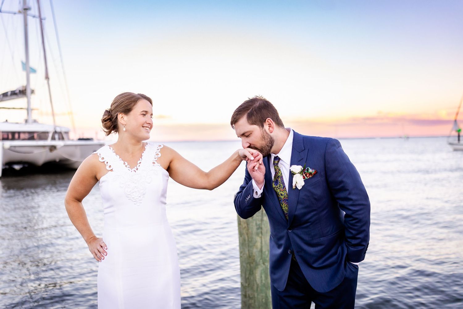 Groom in navy suit kisses bride's hand near waterfront dock at sunset during romantic outdoor wedding ceremony.