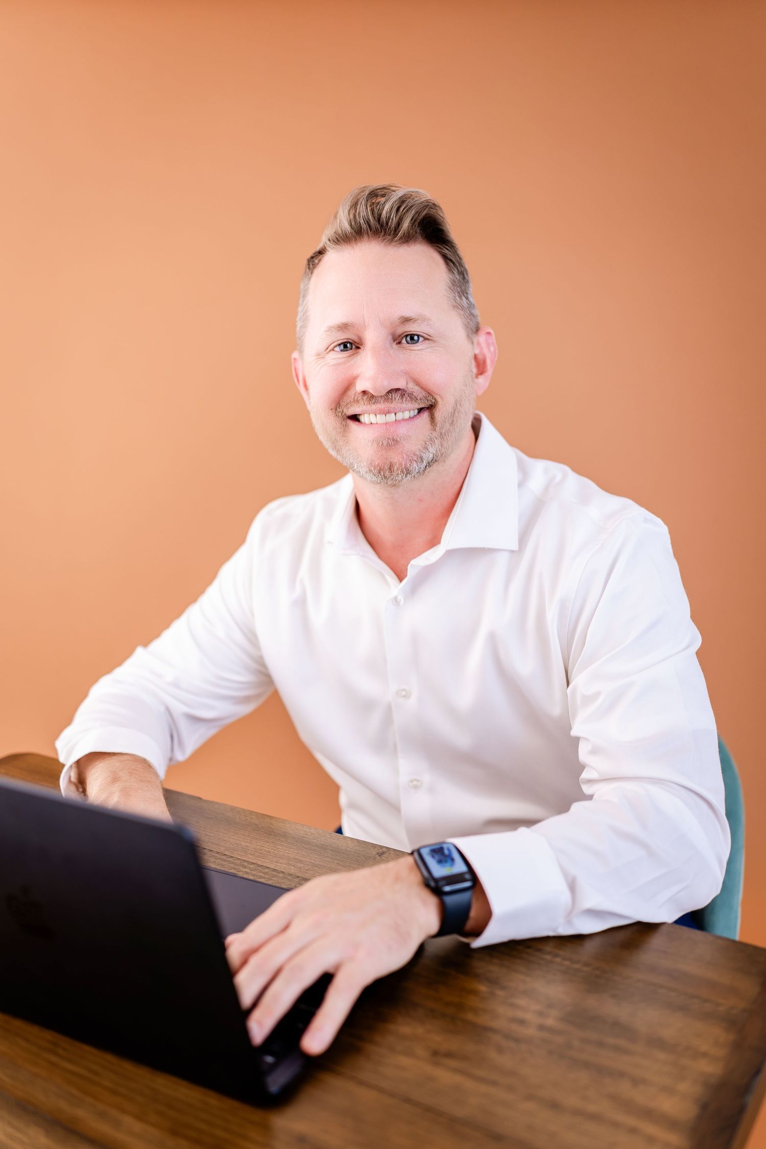 Smiling man in white shirt working on laptop at wooden desk against orange background.