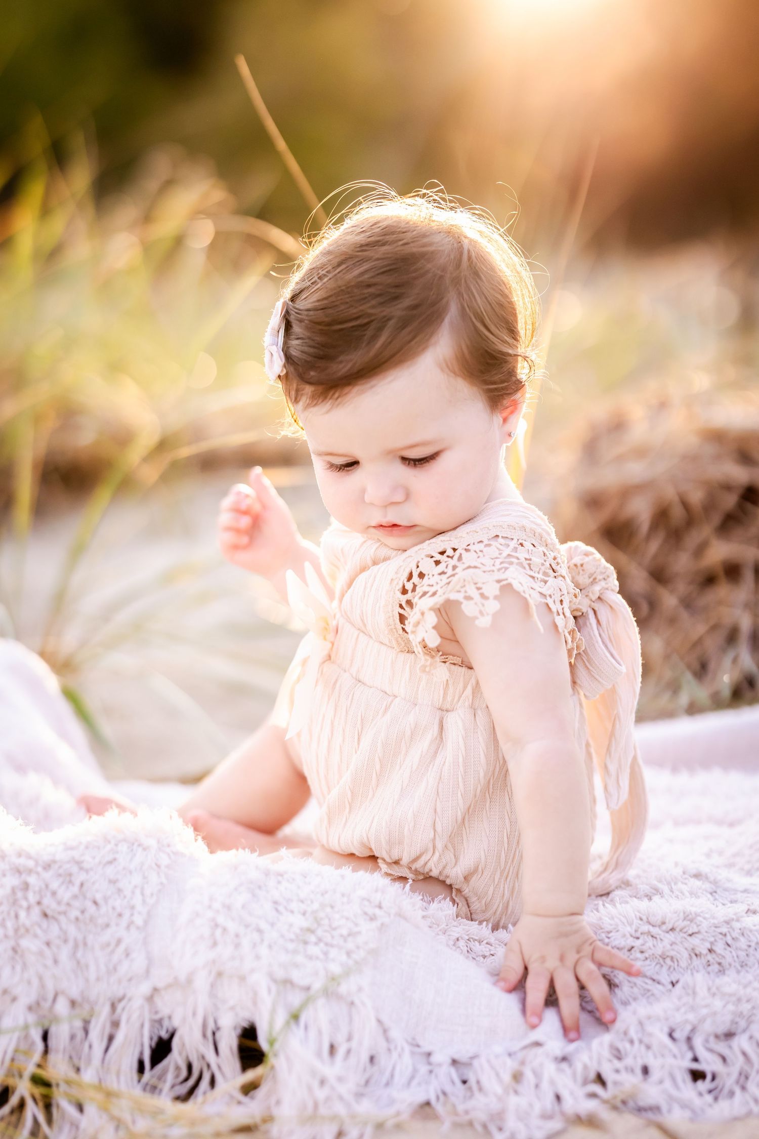 A baby in a pink lace dress lies on a white lace blanket in a dreamy outdoor setting during golden hour.