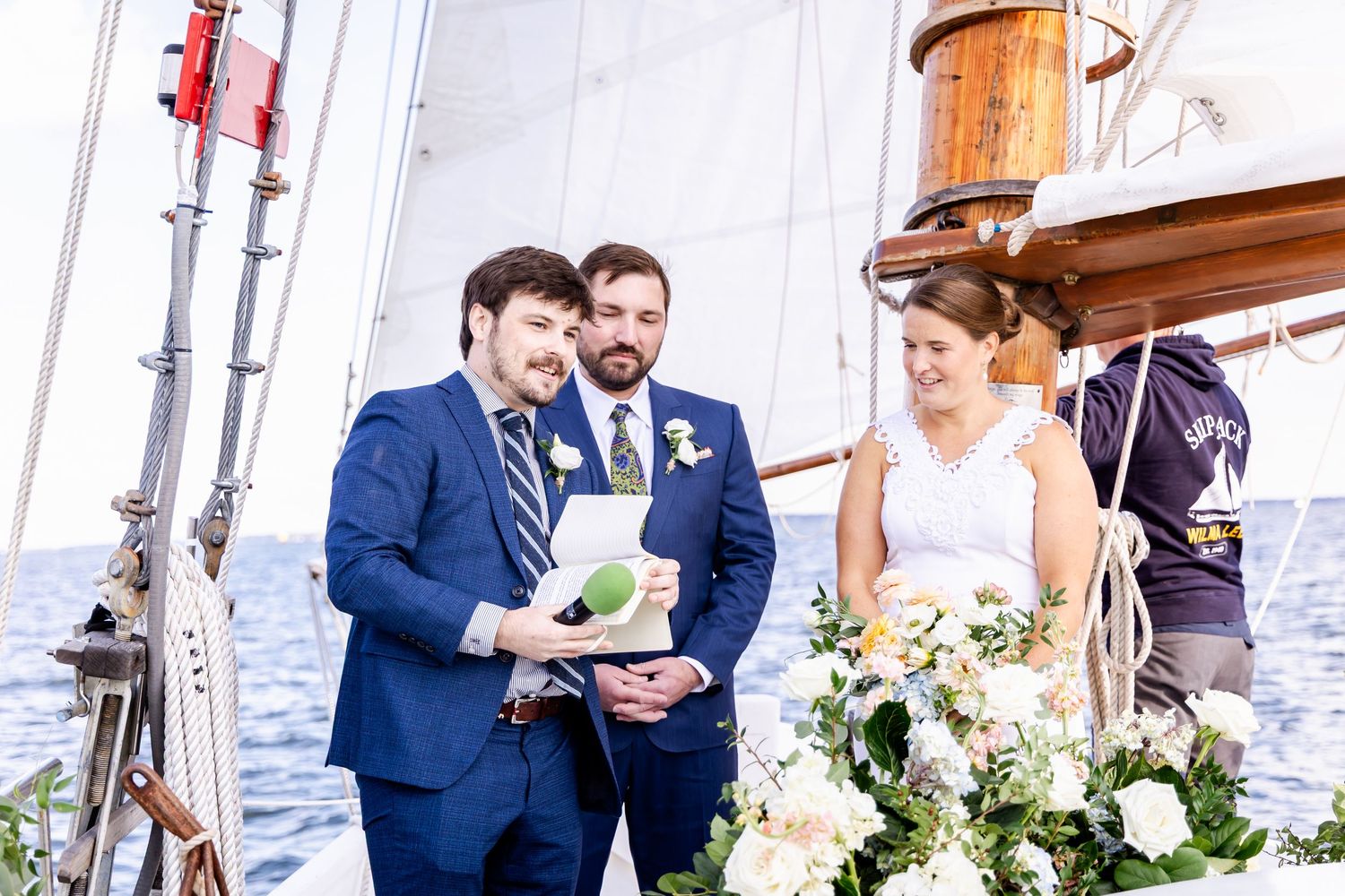 Couple exchanging vows on a sailboat wedding ceremony with floral arrangements and officiant on blue water.