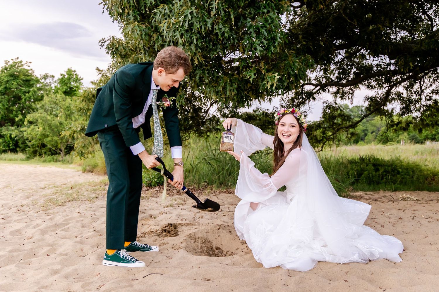 A couple in wedding attire playfully pose together on a sandy beach path surrounded by greenery.