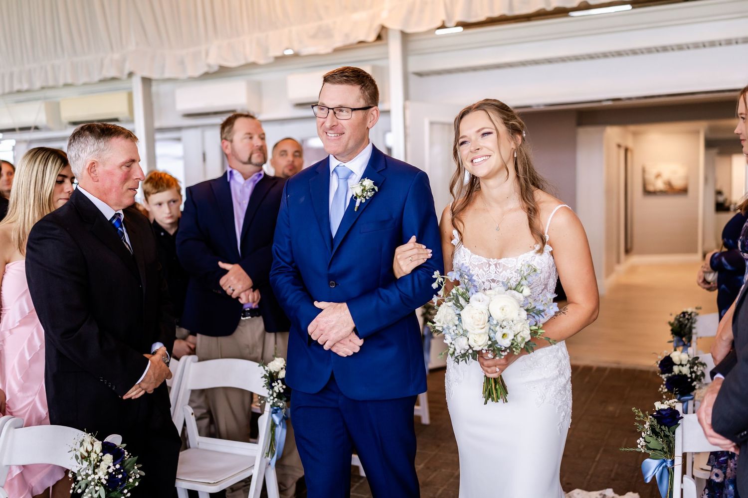 A couple stands at the altar during their wedding ceremony in an elegant indoor venue with white chairs and floral decor.