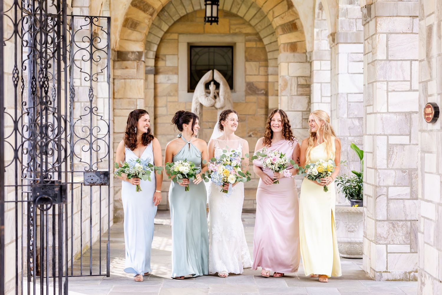 A wedding party in pastel dresses stands in front of a stone church with wrought iron gates and floral decorations.