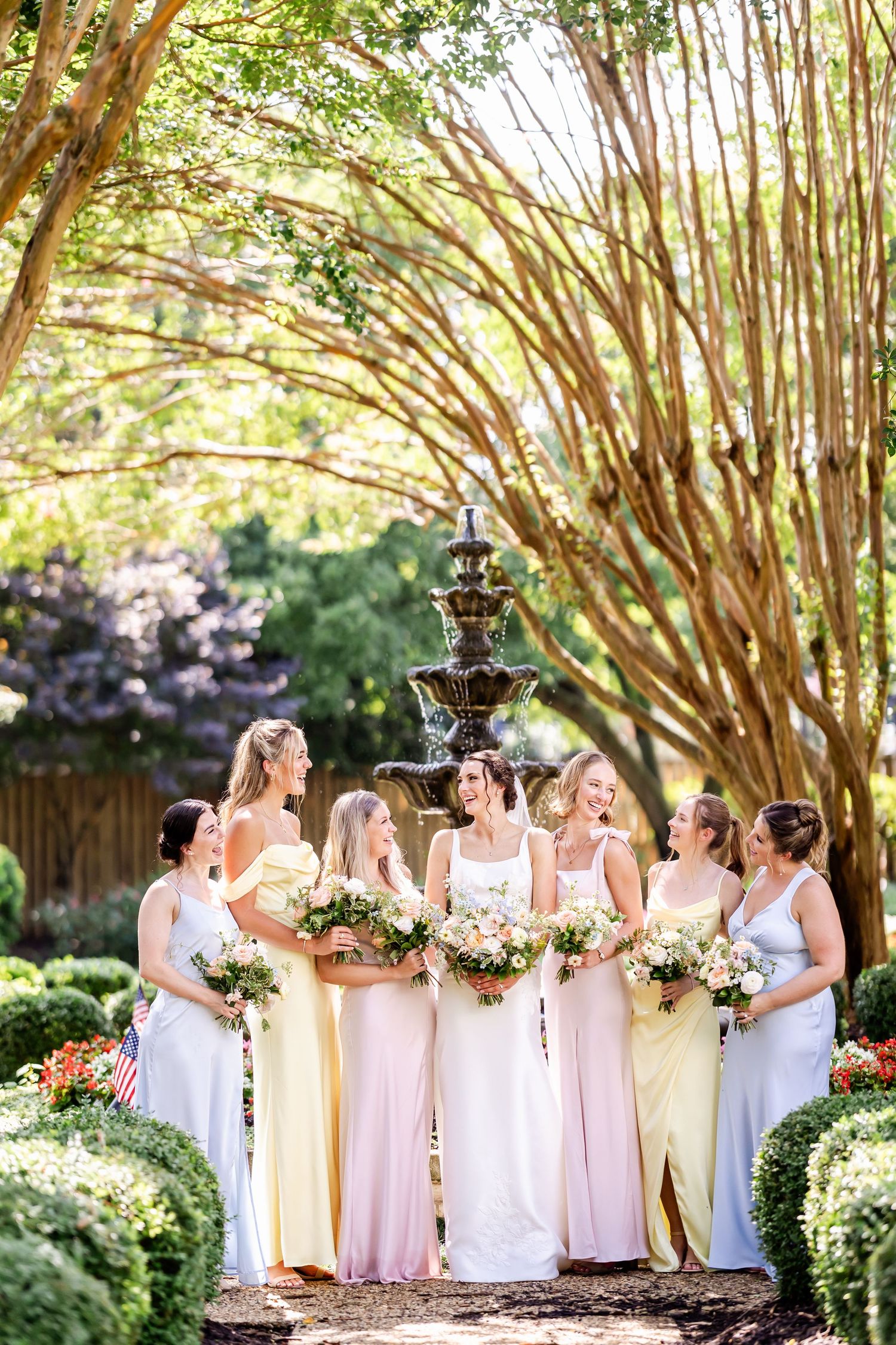 Bridesmaids in pastel dresses gather around fountain holding bouquets in sunlit garden.