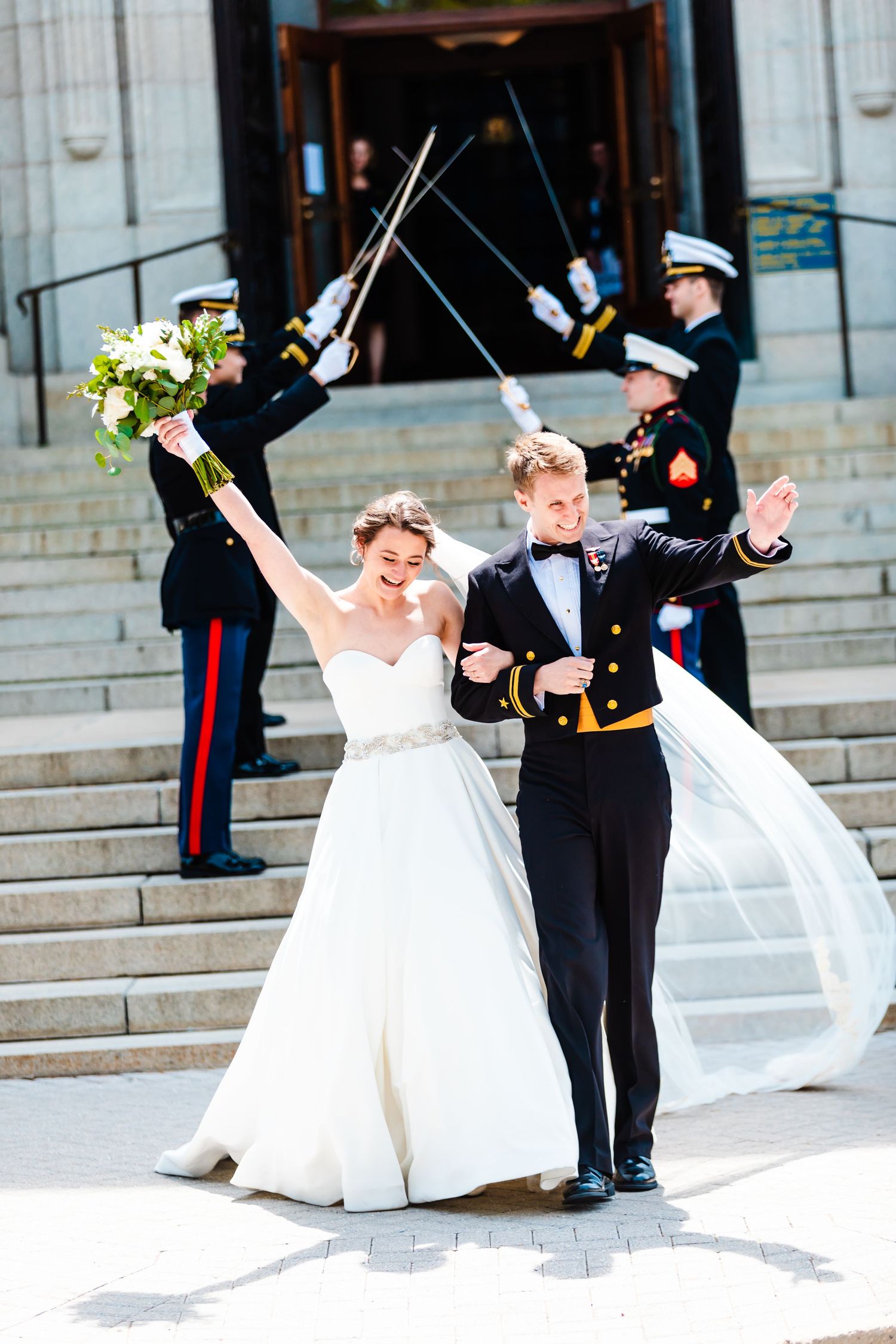 Bride and groom in military dress uniform celebrate with playful poses on stone steps during their wedding ceremony.