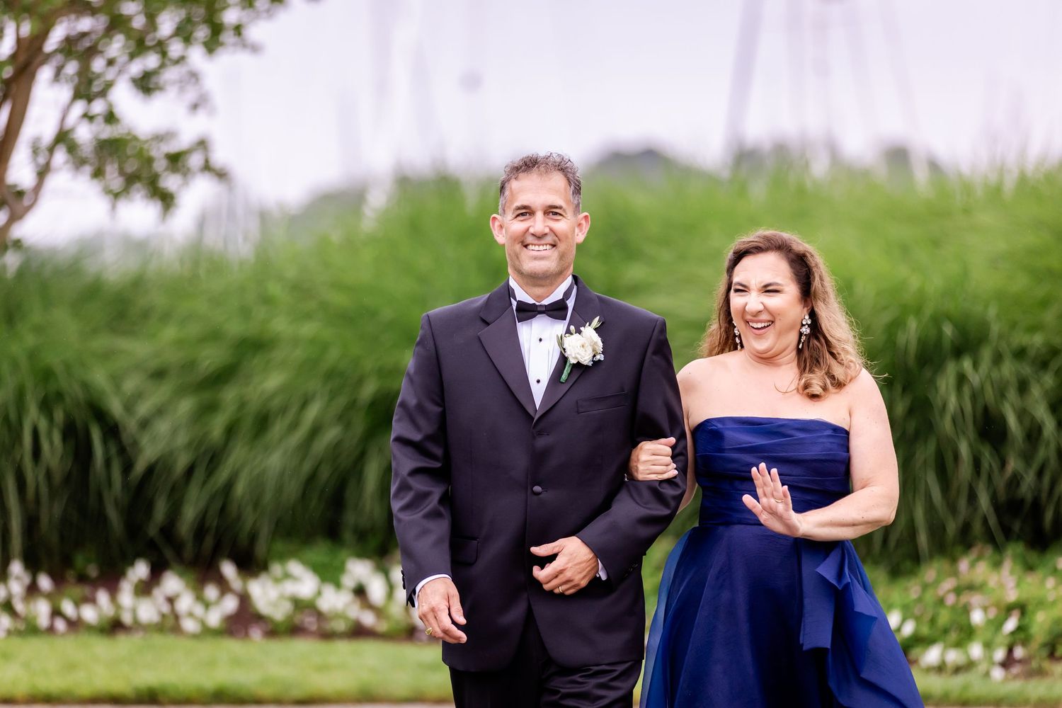 A pair in formal attire smile together at an outdoor event with tall grasses in the background.