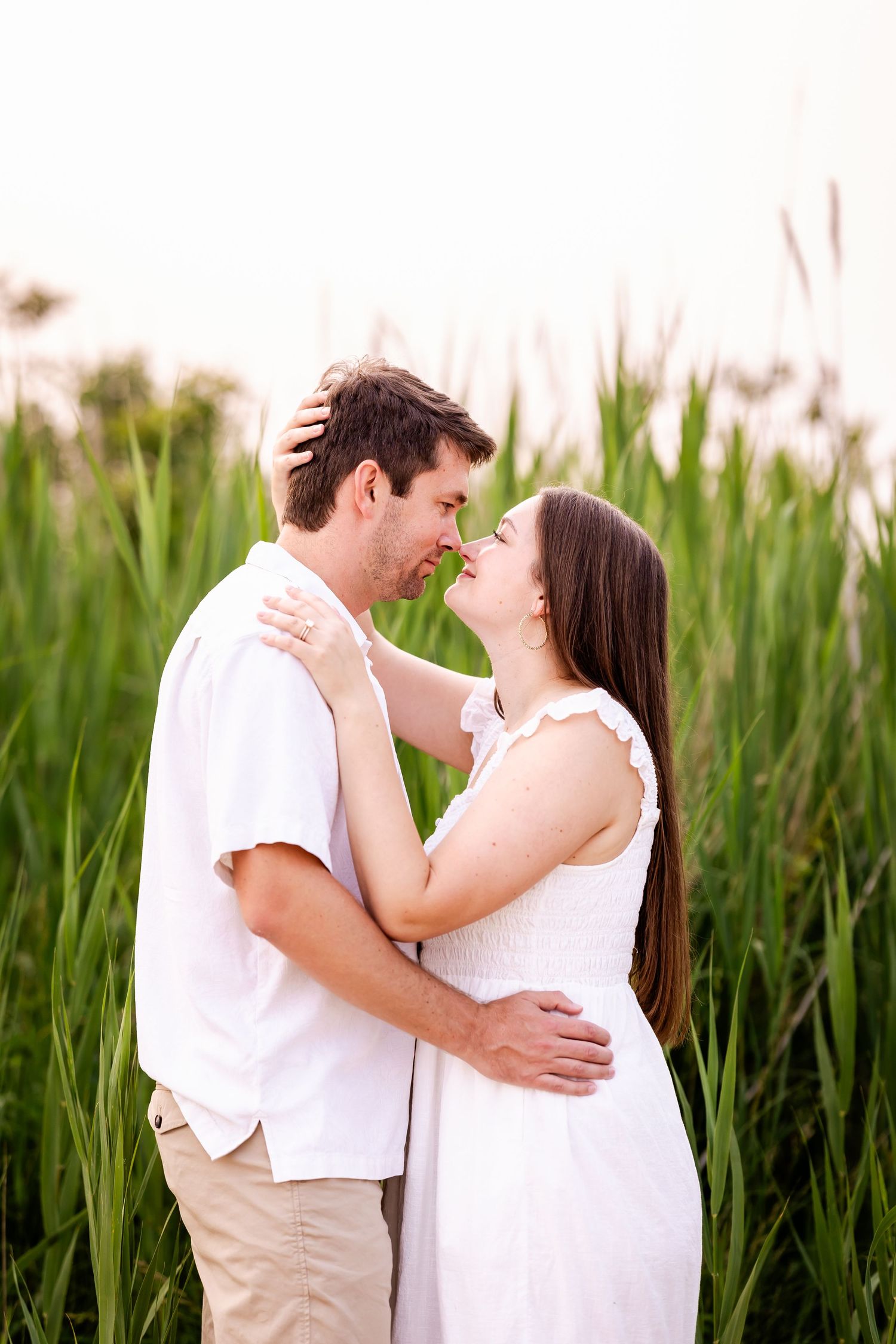 A romantic couple in white outfits shares tender moments in a field of tall green grass.
