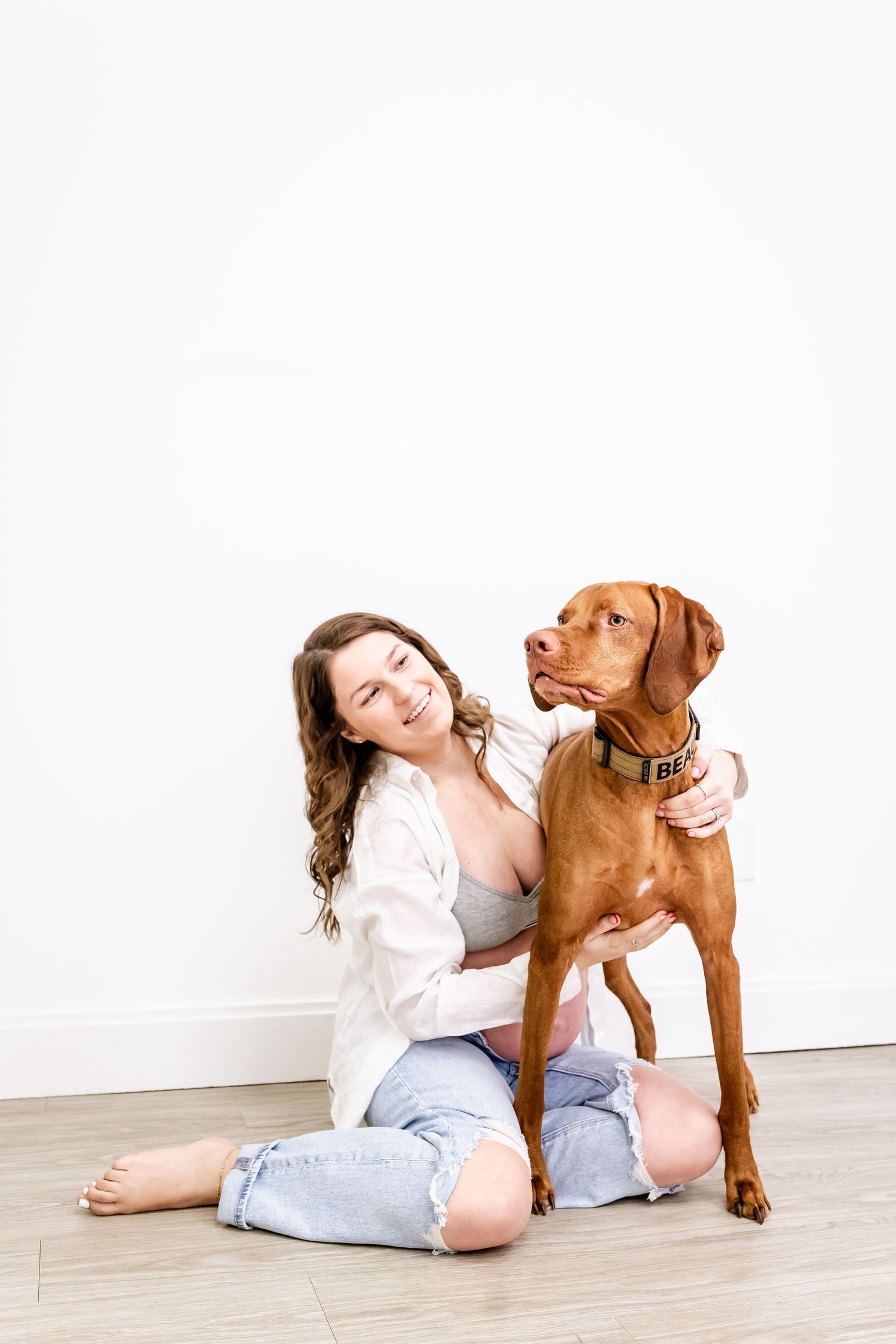A Vizsla dog sits beside a person in casual clothing on a light wooden floor against a white wall.