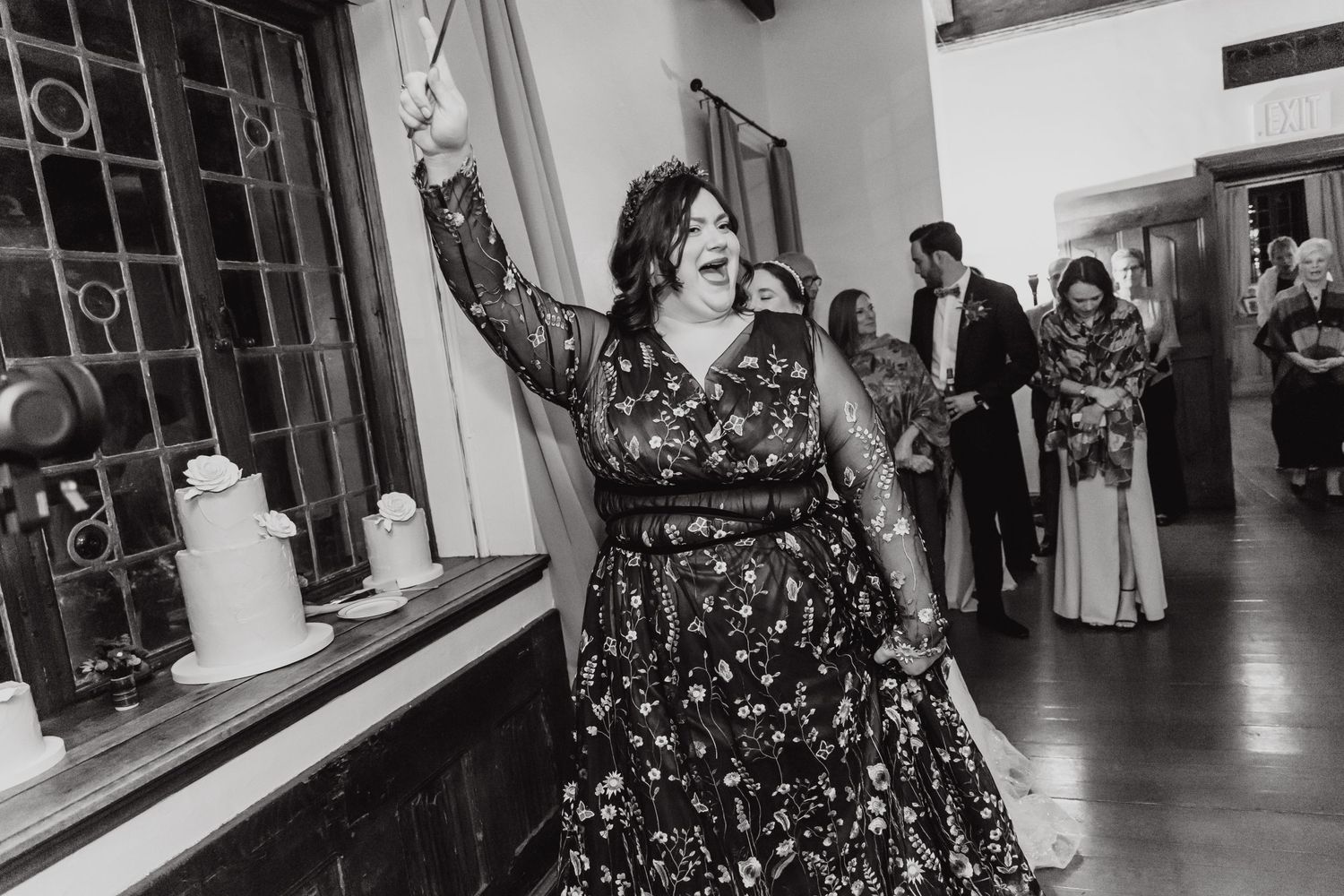 Person in floral dress dances joyfully at indoor reception while guests mingle in background.
