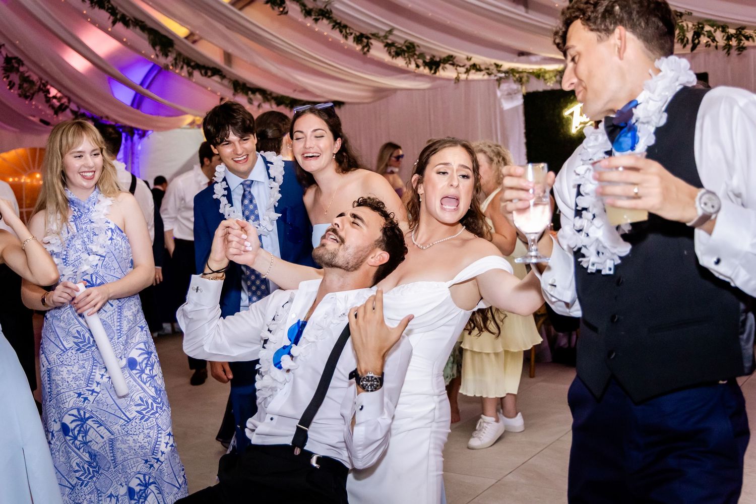 Wedding reception guests dance and celebrate under a draped tent ceiling with festive lighting.