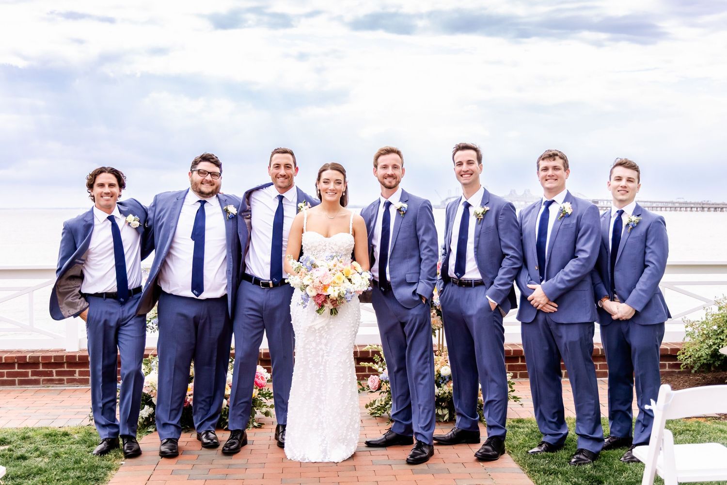 Wedding party in navy suits and white dress lined up for formal photos by the water.