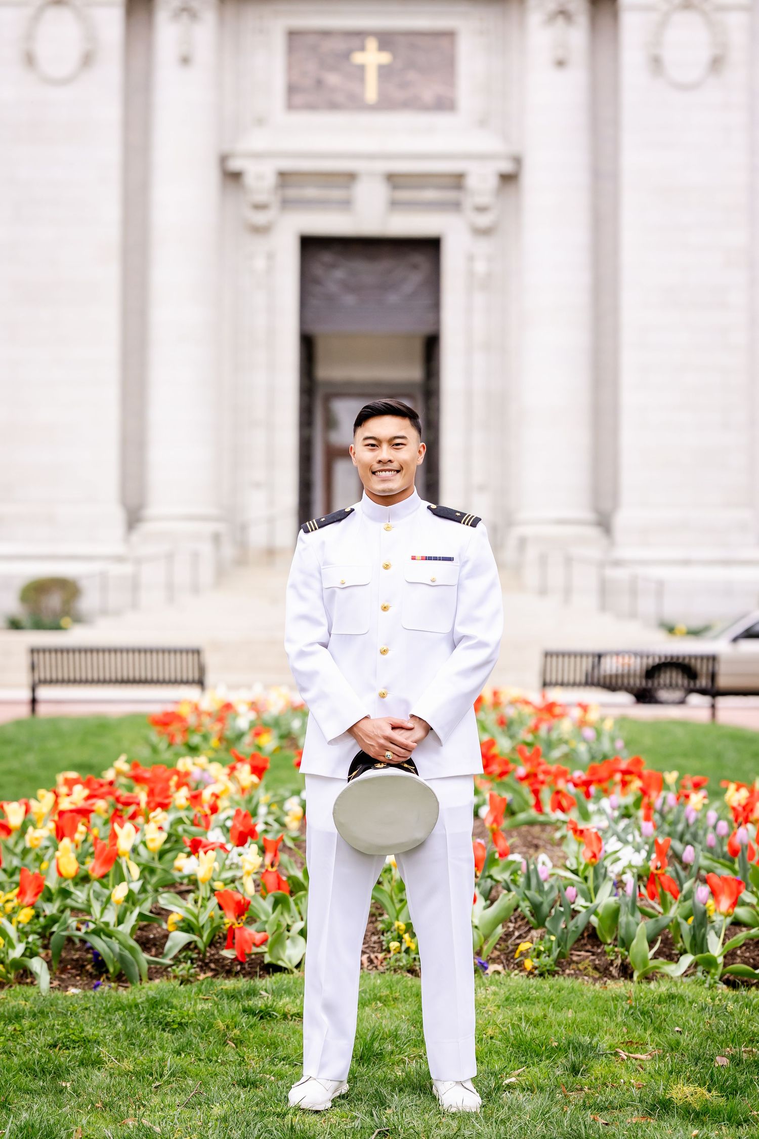 Military service member standing in formal white uniform in front of church entrance with blooming tulip garden.