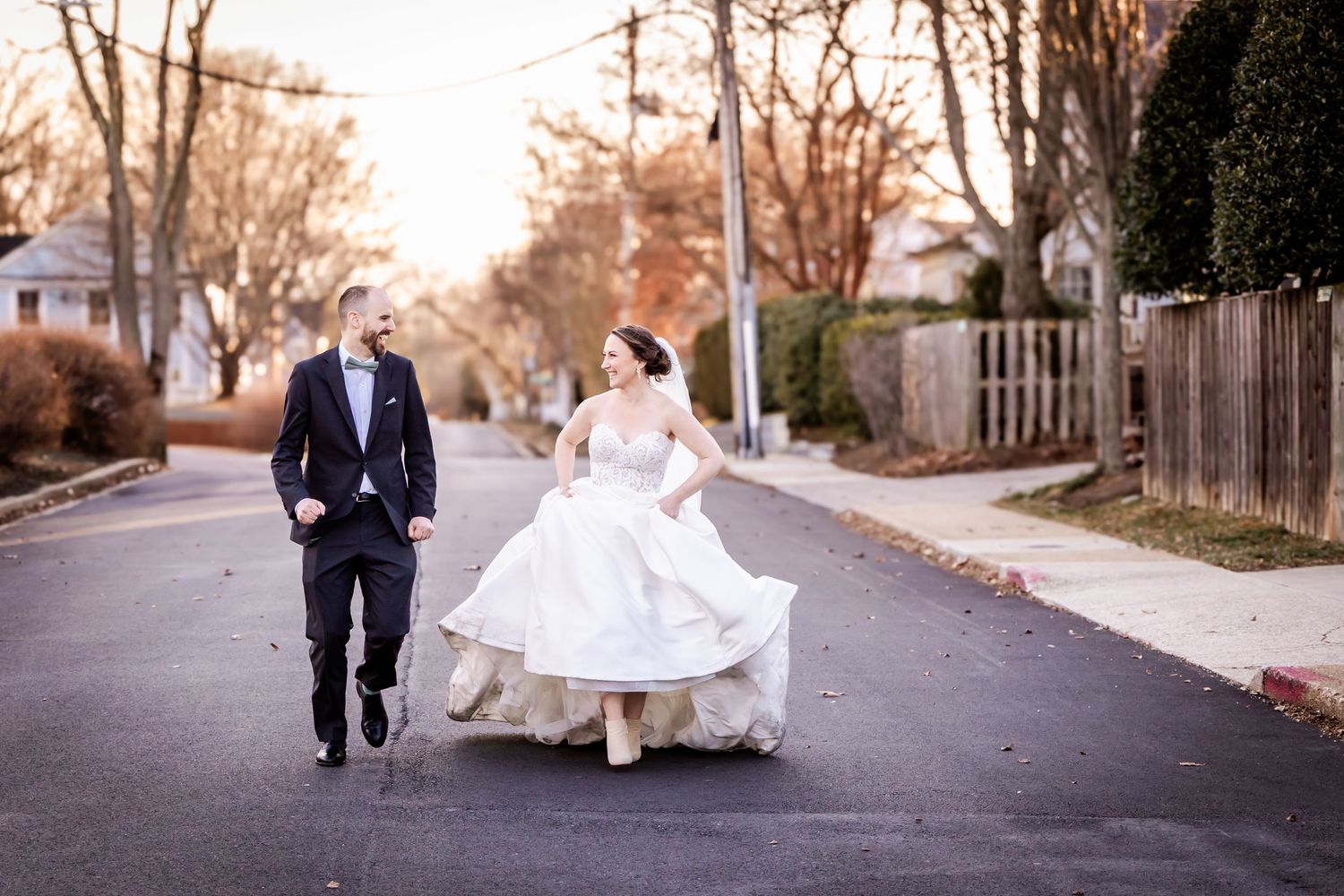 Two people walk down a tree-lined residential street at sunset, with one in a flowing white gown.