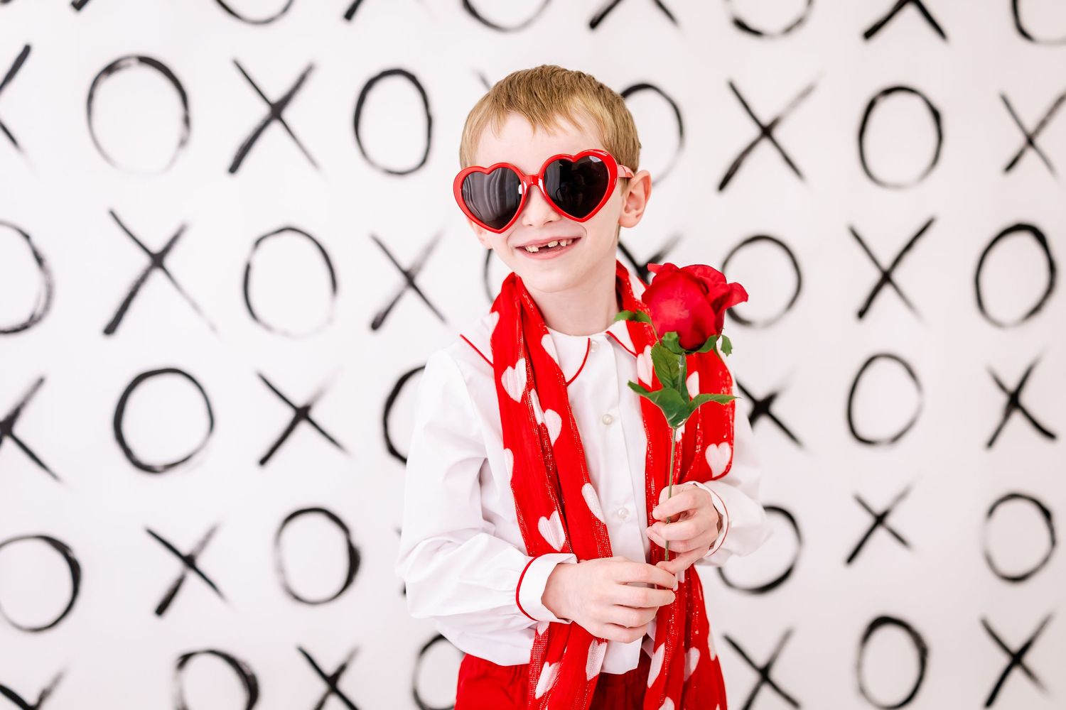 Subject wearing heart-shaped sunglasses and red vest holds red roses against black and white XO pattern background.