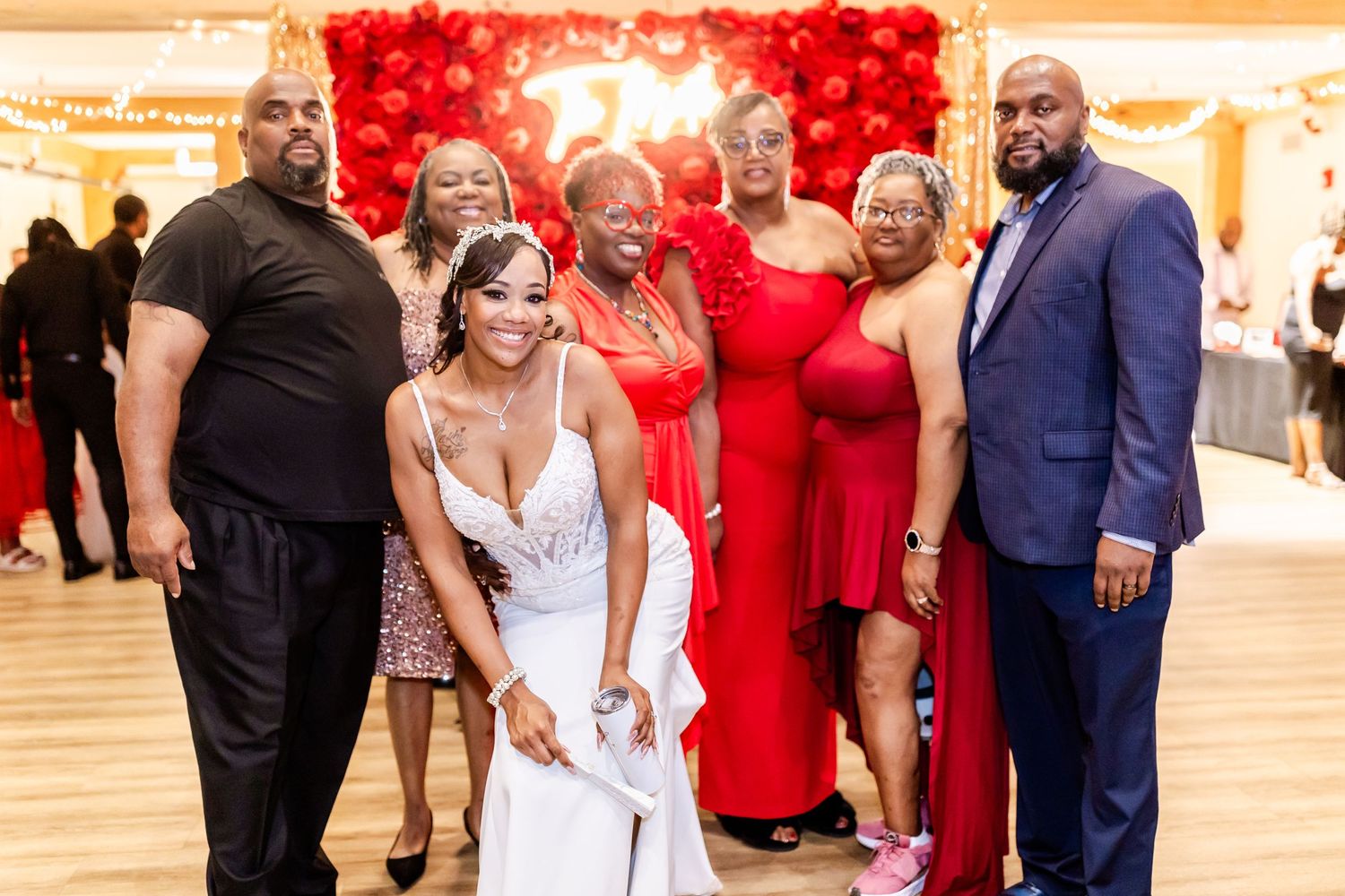 Wedding party members posing together in red and formal attire against a rose wall backdrop.