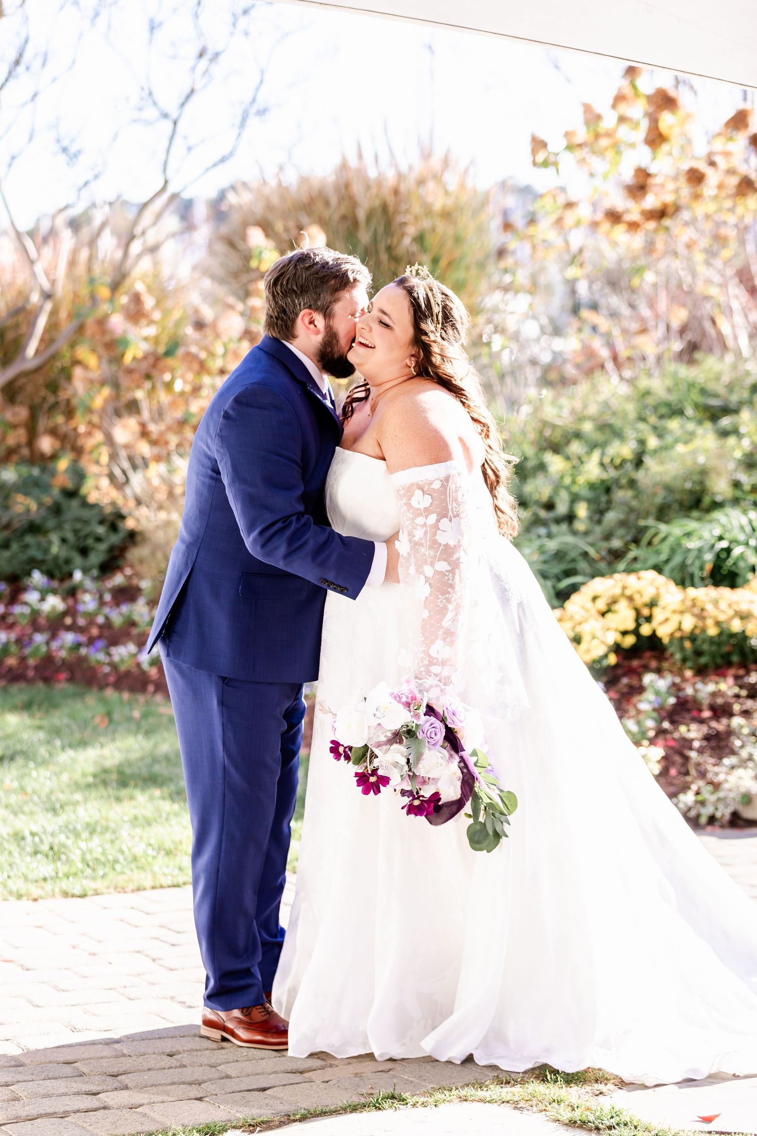 Romantic wedding couple embracing in outdoor garden setting with purple floral bouquet.