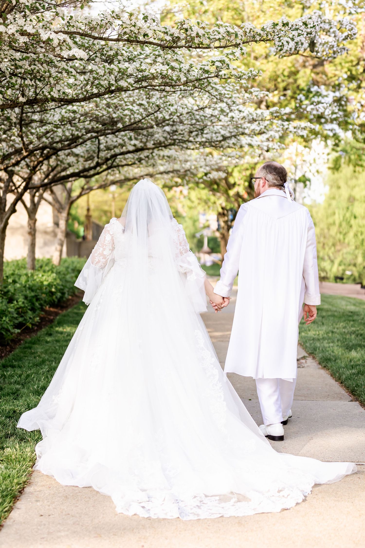 A couple in white wedding attire walk down a path under blooming cherry blossom trees.