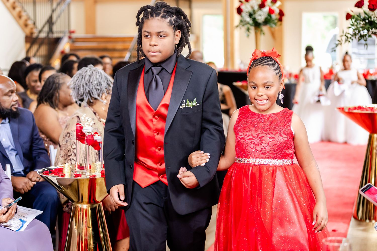 Wedding party members walk down the red carpet aisle in coordinated black and red formal attire.