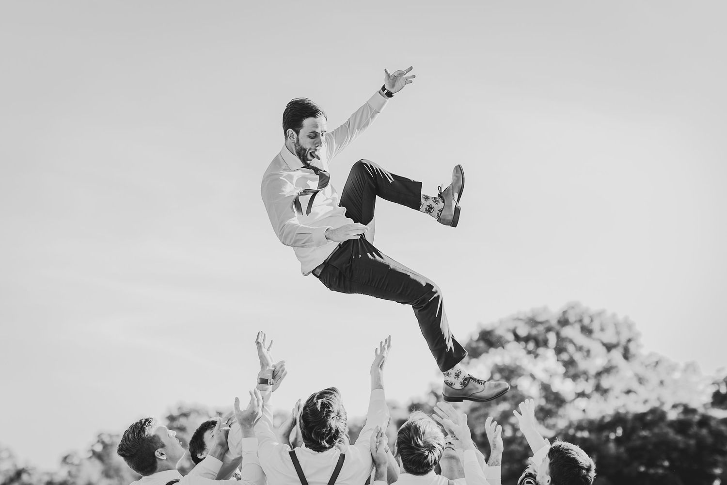 A crowd enthusiastically tosses a person high into the air during an energetic outdoor celebration in black and white.