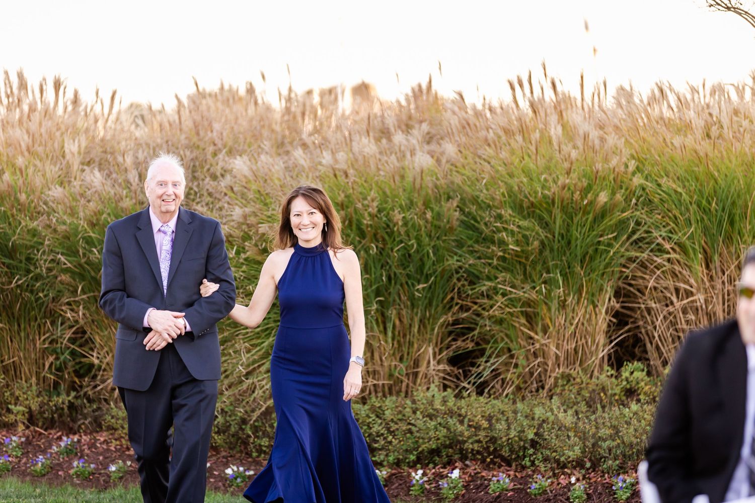 Two elegantly dressed attendants walk down a garden path with onlookers visible.