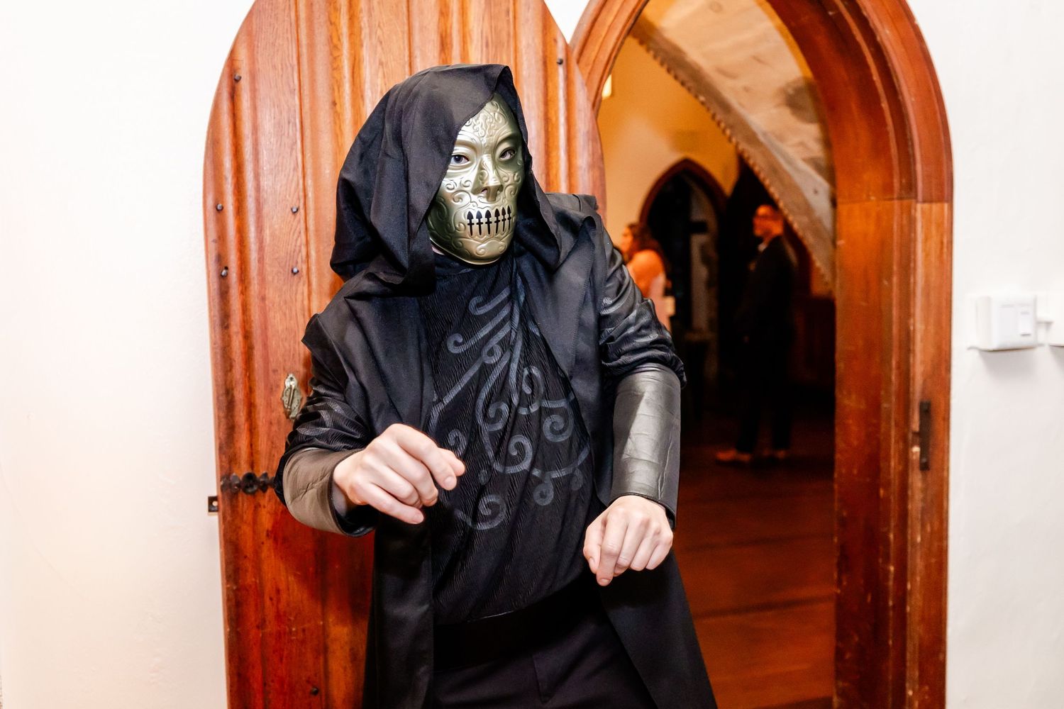 Person wearing spooky skeleton mask and black hooded costume poses in arched doorway.