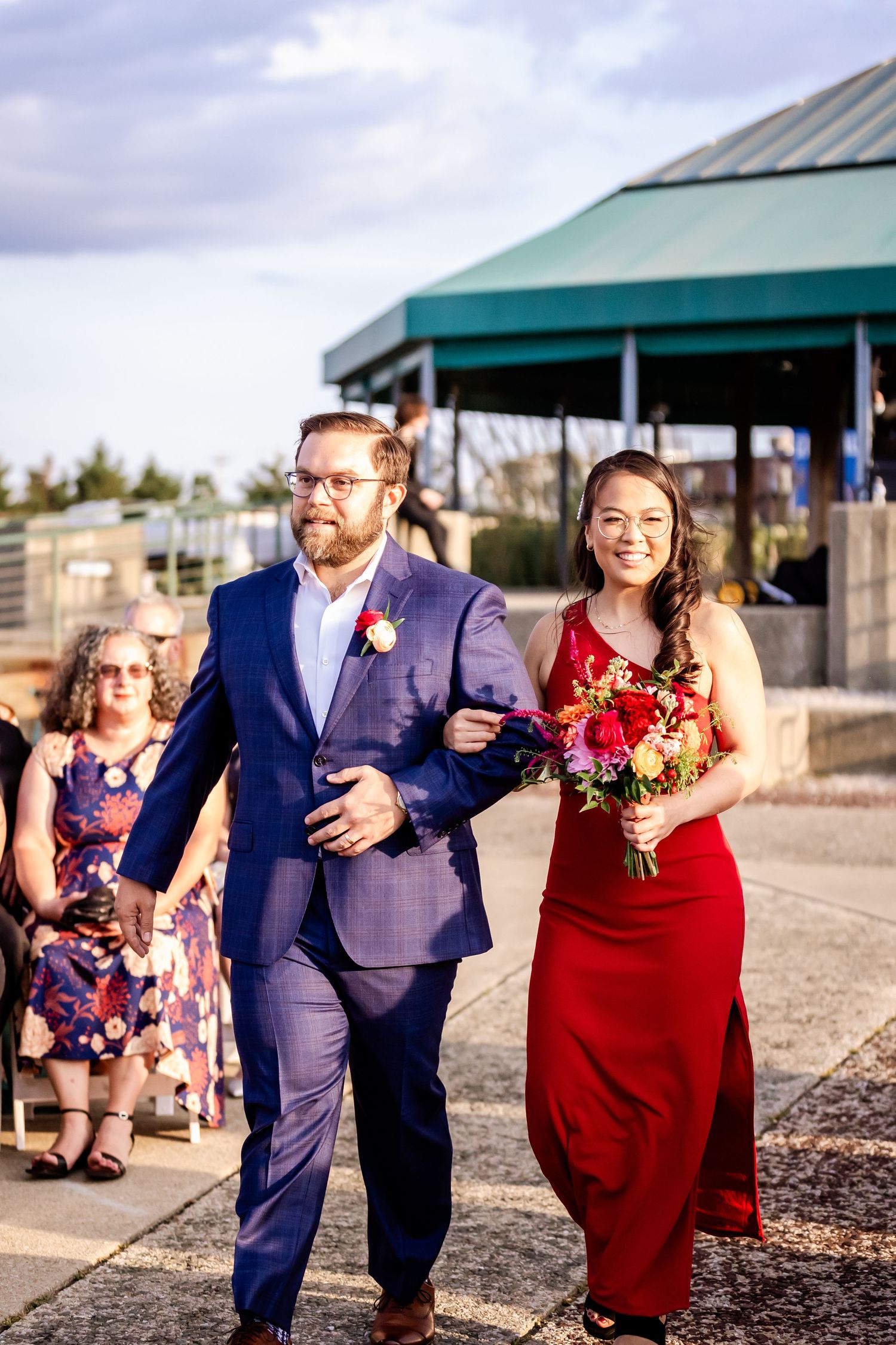 Wedding party members walk along a waterfront path with a woman in a red dress holding a vibrant bouquet.