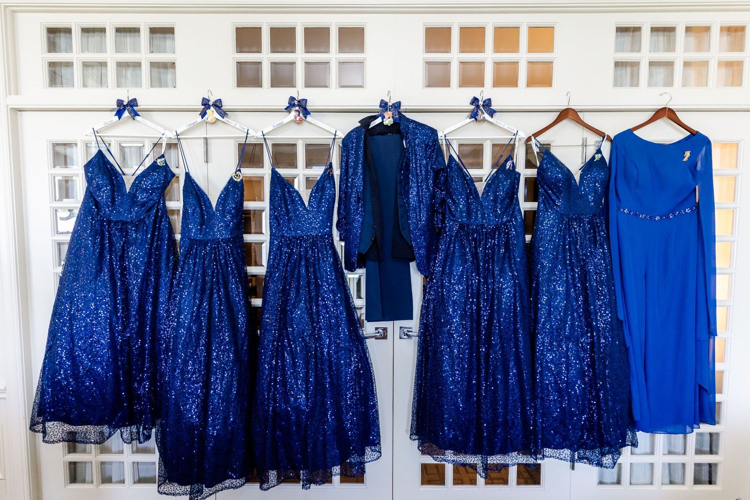 Sparkly royal blue bridesmaid dresses hanging in a row on white hangers against a window background.