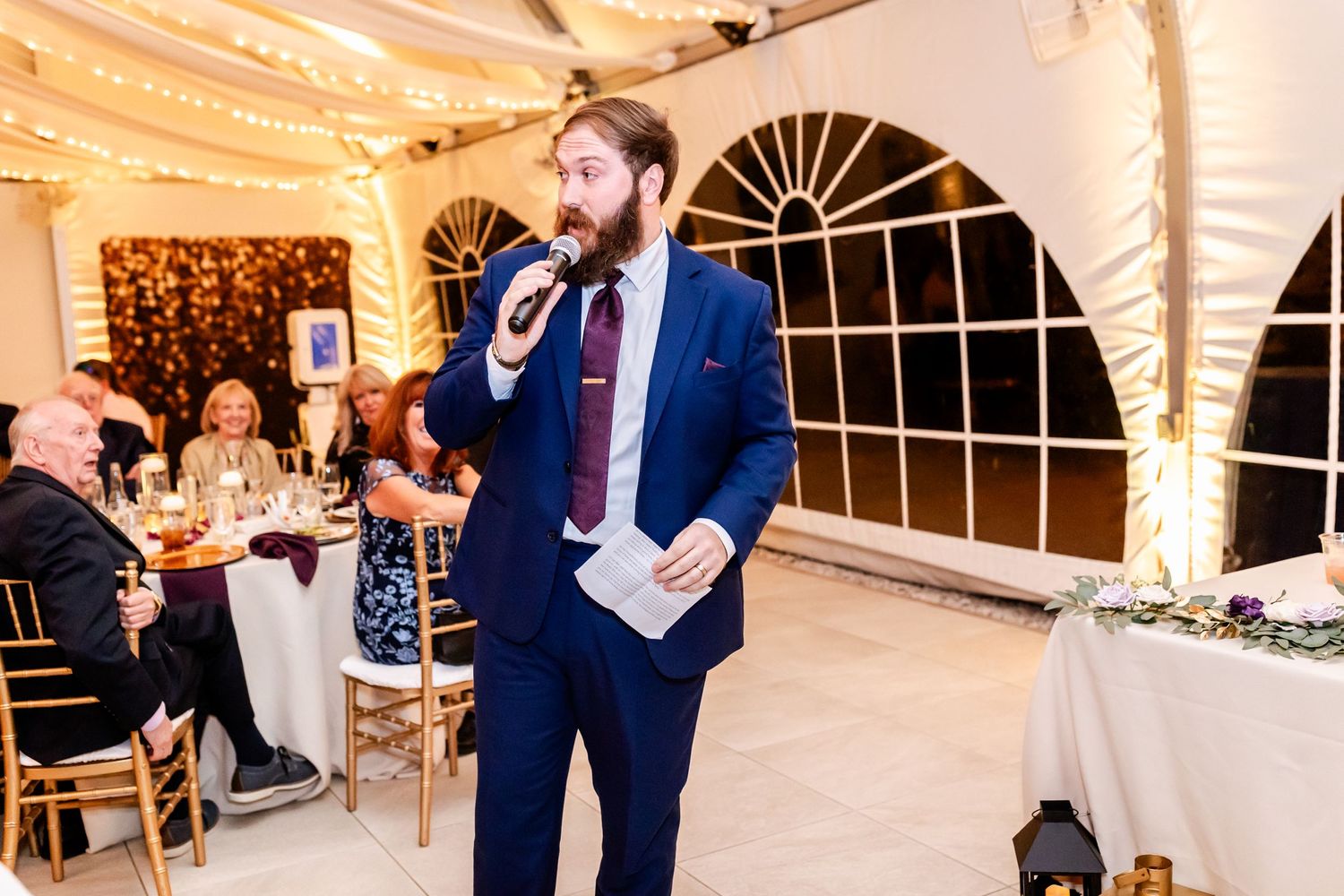 Wedding guest giving speech during reception in decorated tent venue with string lights.