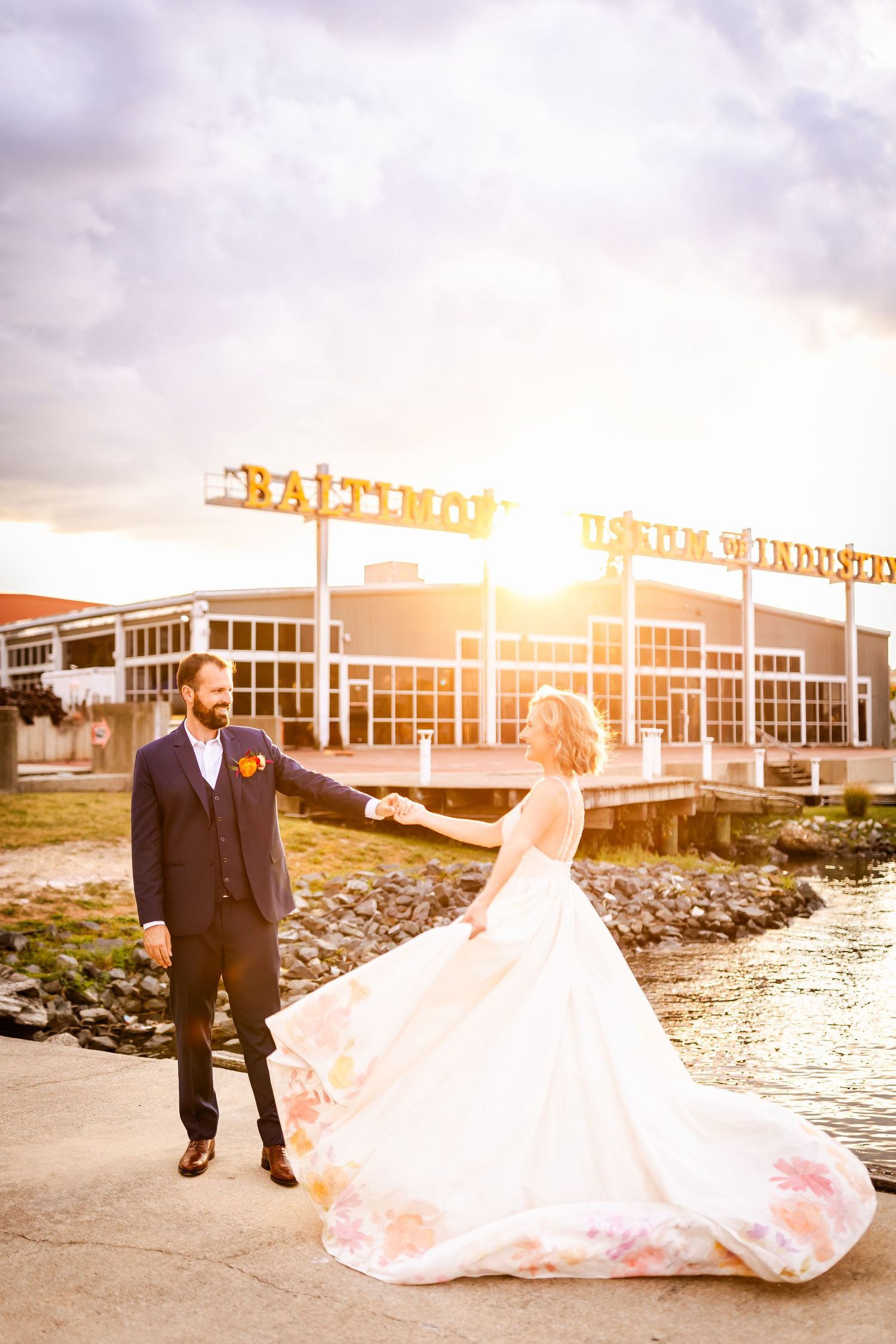 A couple in wedding attire poses near a waterfront at golden hour with industrial buildings in the background.