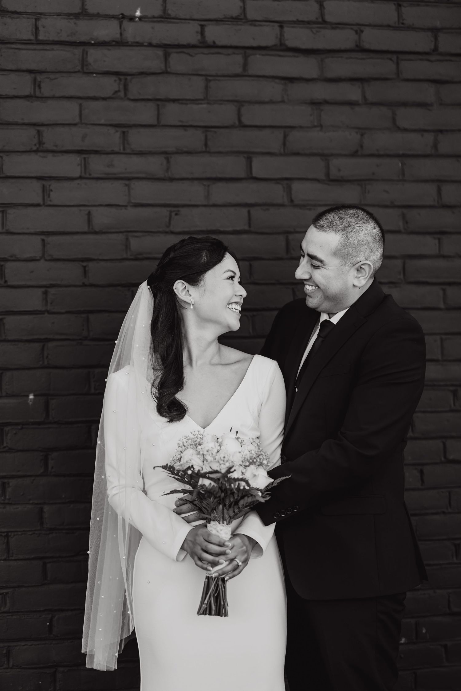 Black and white portrait of newlyweds against brick wall with bride holding bouquet.