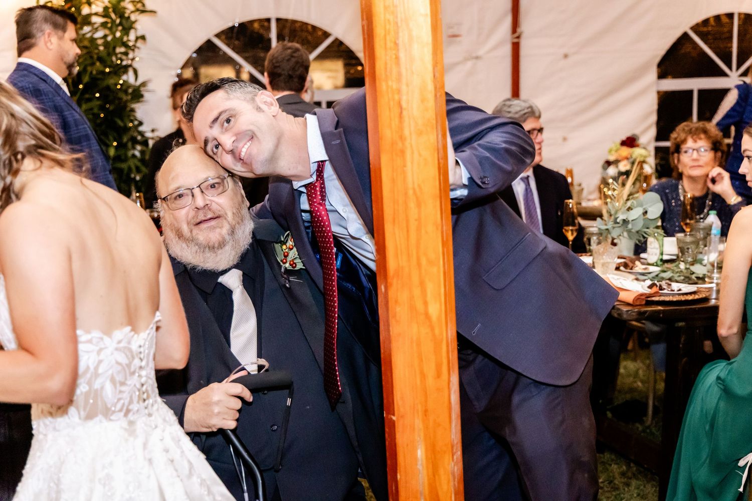 Guests celebrating at a lively wedding reception under a white tent with warm lighting and wooden beams.
