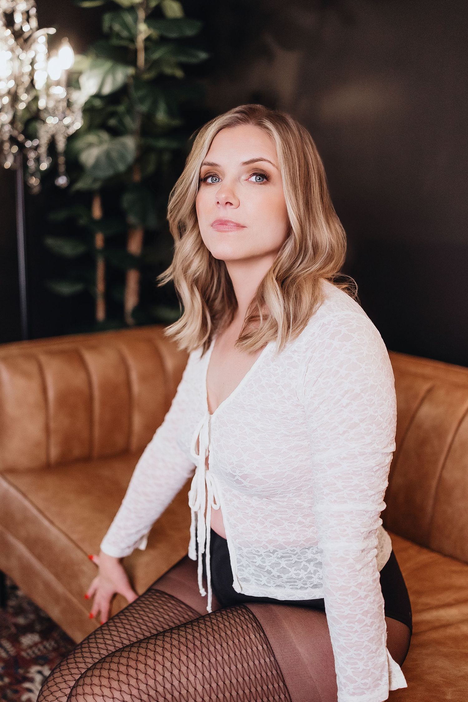 A woman in a white lace top and fishnet stockings poses on a brown leather couch in an elegant setting.