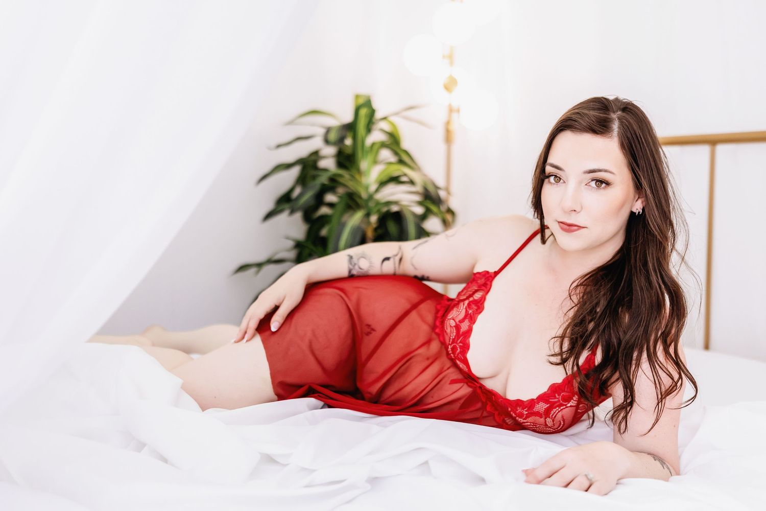 Woman in sheer red lace loungewear reclines gracefully against white pillows in bedroom setting.