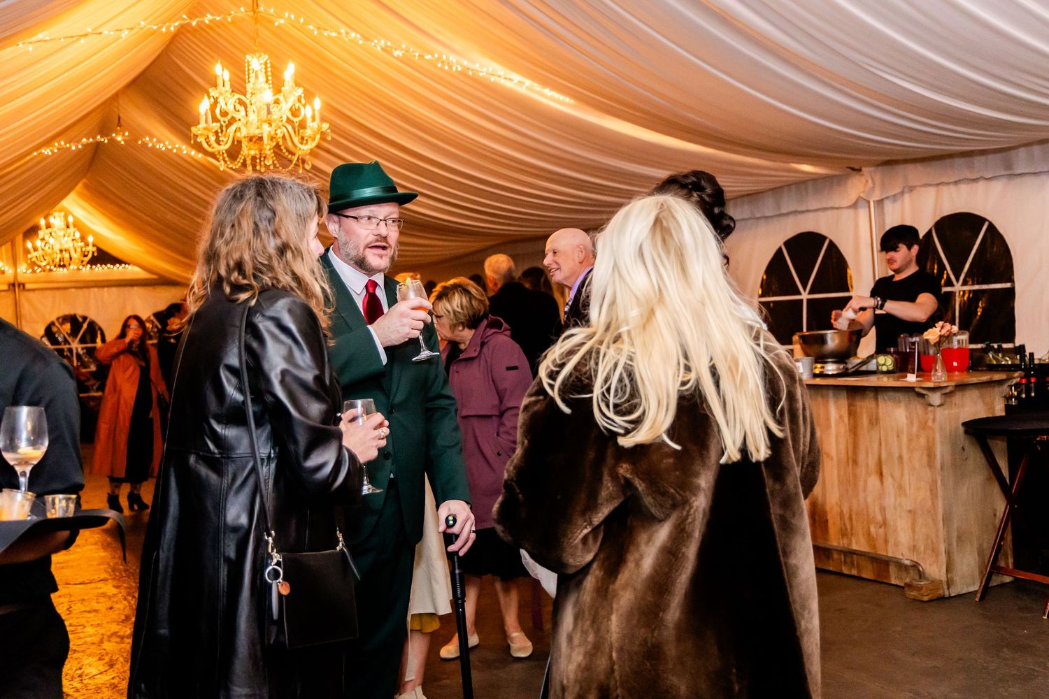 Wedding reception guests mingling under a warmly lit tent with chandeliers.