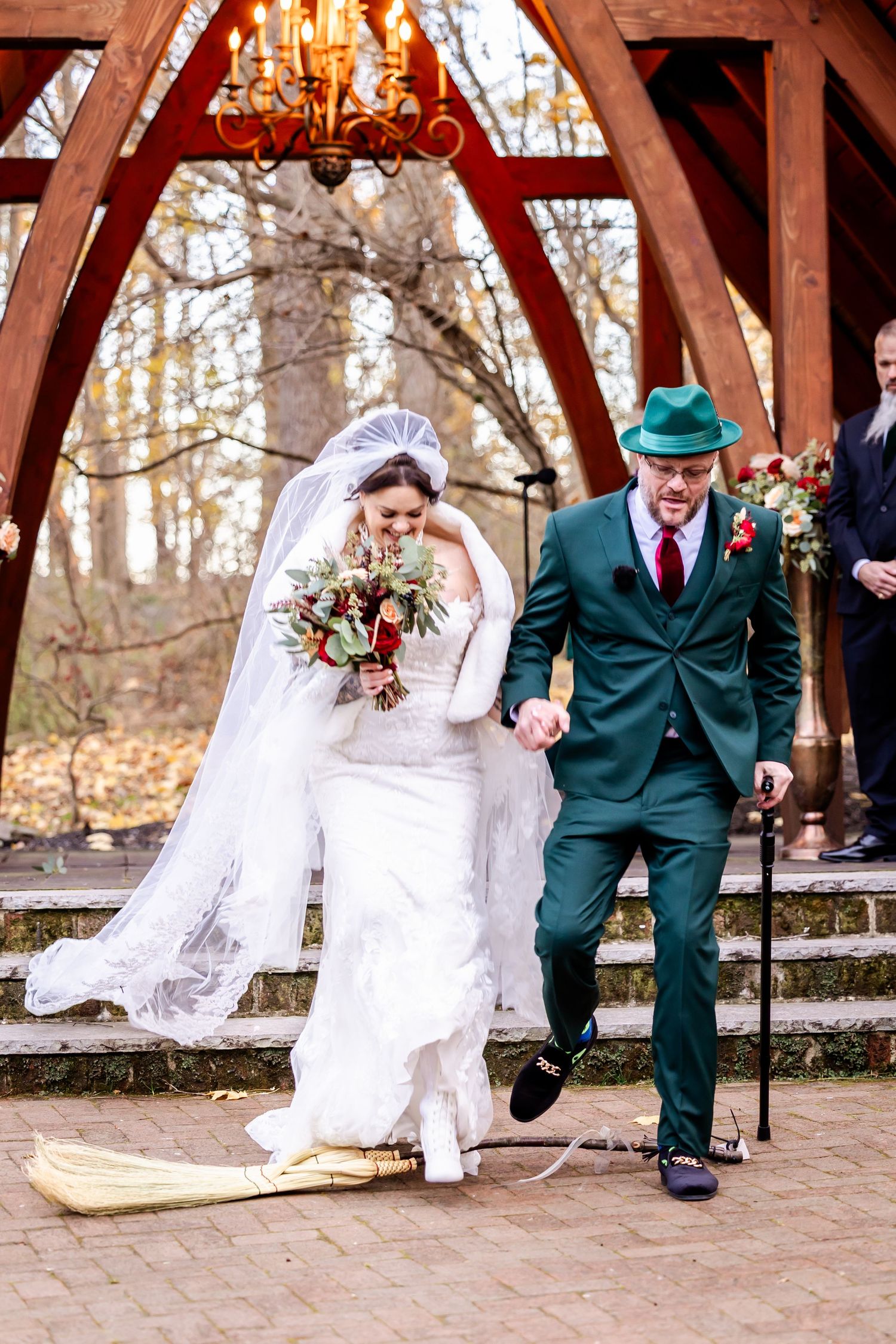 A couple walks under a wooden arch during their wedding ceremony with the groom in a green suit.