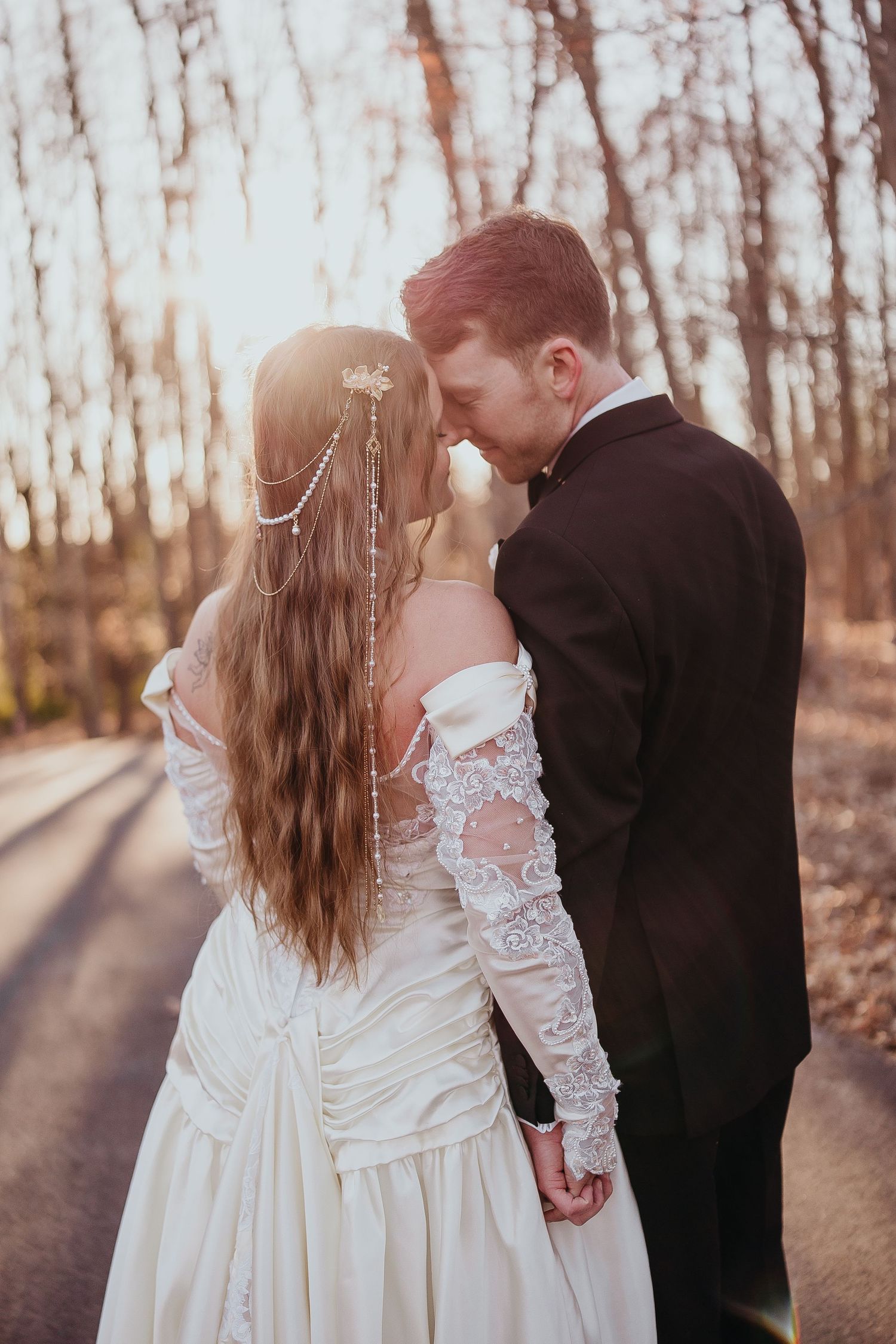 Bride and groom sharing a romantic moment on a sunlit forest road, bride in lace wedding gown holding hands.