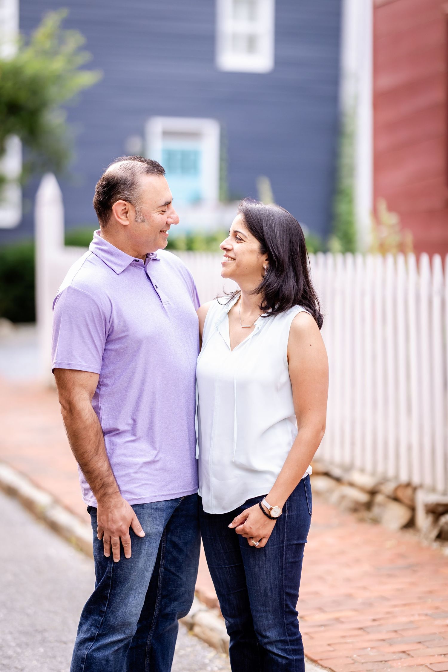 A couple shares a joyful moment laughing together outdoors near a white picket fence.
