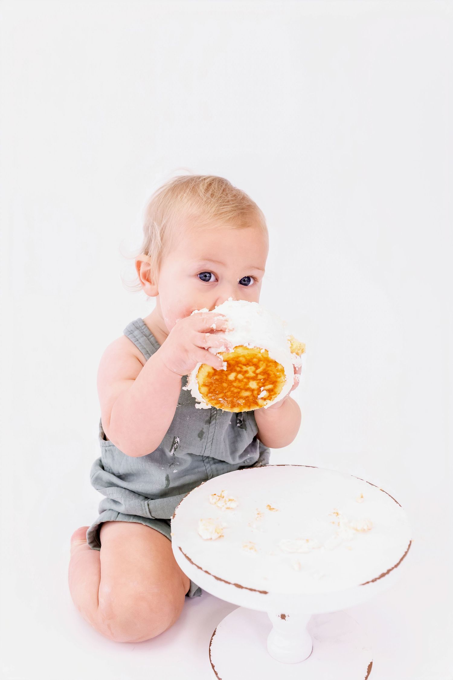 Baby in gray onesie eagerly eats orange food from white plate while sitting on white background.