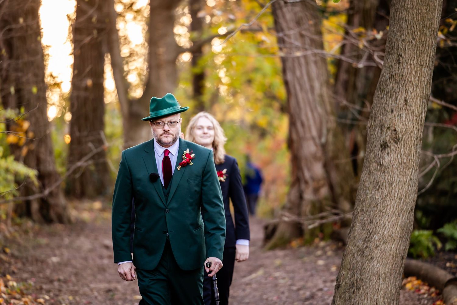 A couple walking through an autumn forest with golden foliage and warm sunlight filtering through the trees.