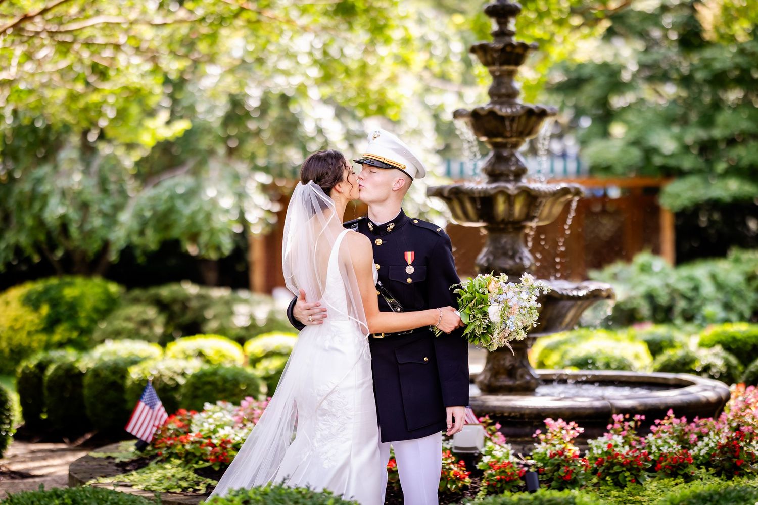 A romantic wedding portrait of a couple embracing by an ornate garden fountain surrounded by blooming flowers.