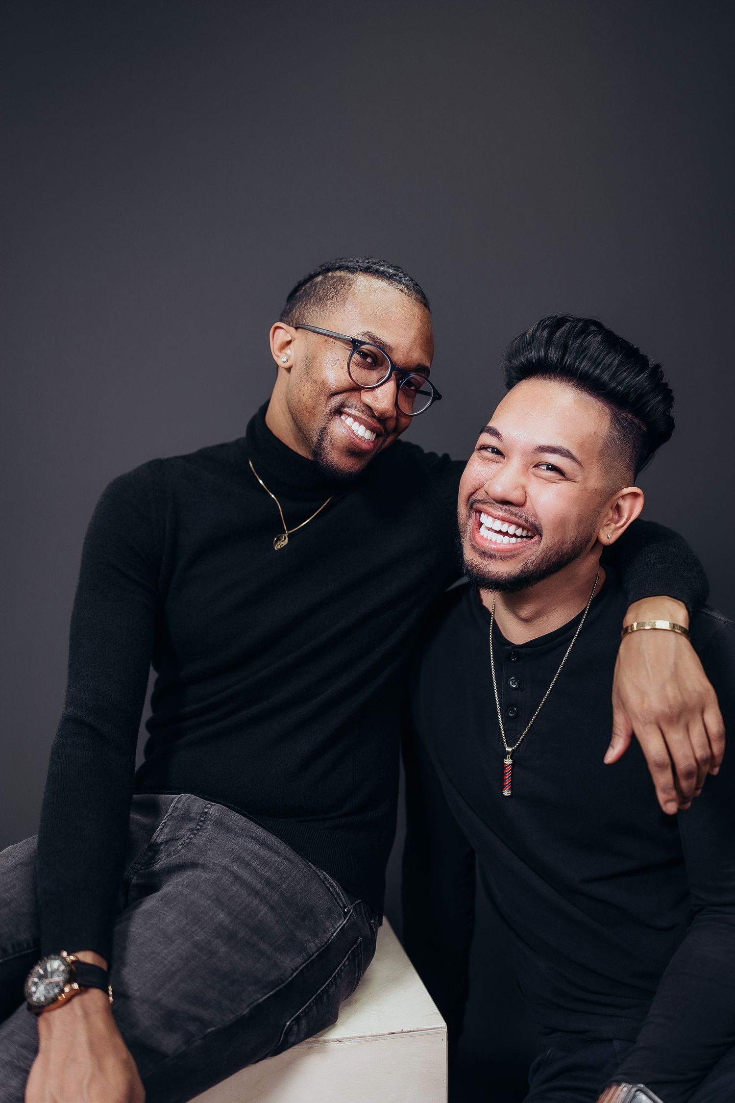 Two friends in black sweaters share genuine laughter and embraces during a casual photo shoot.