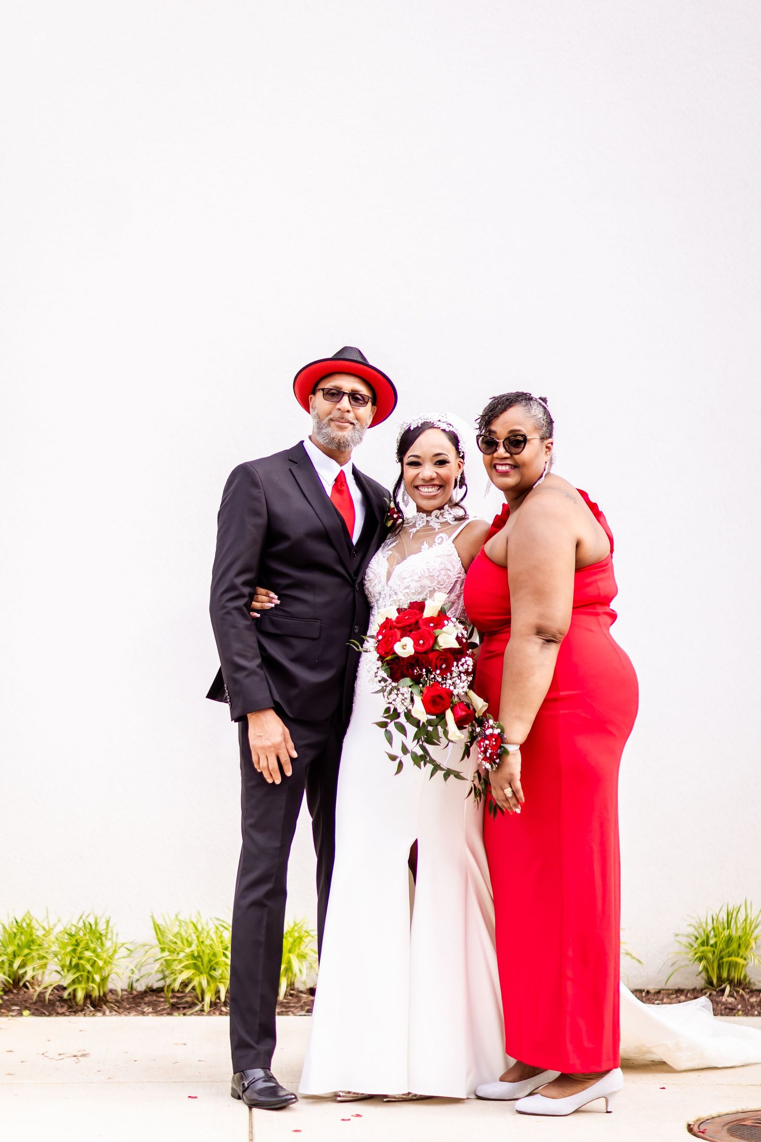Wedding group with black suit, white dress and red gown pose formally against white backdrop.