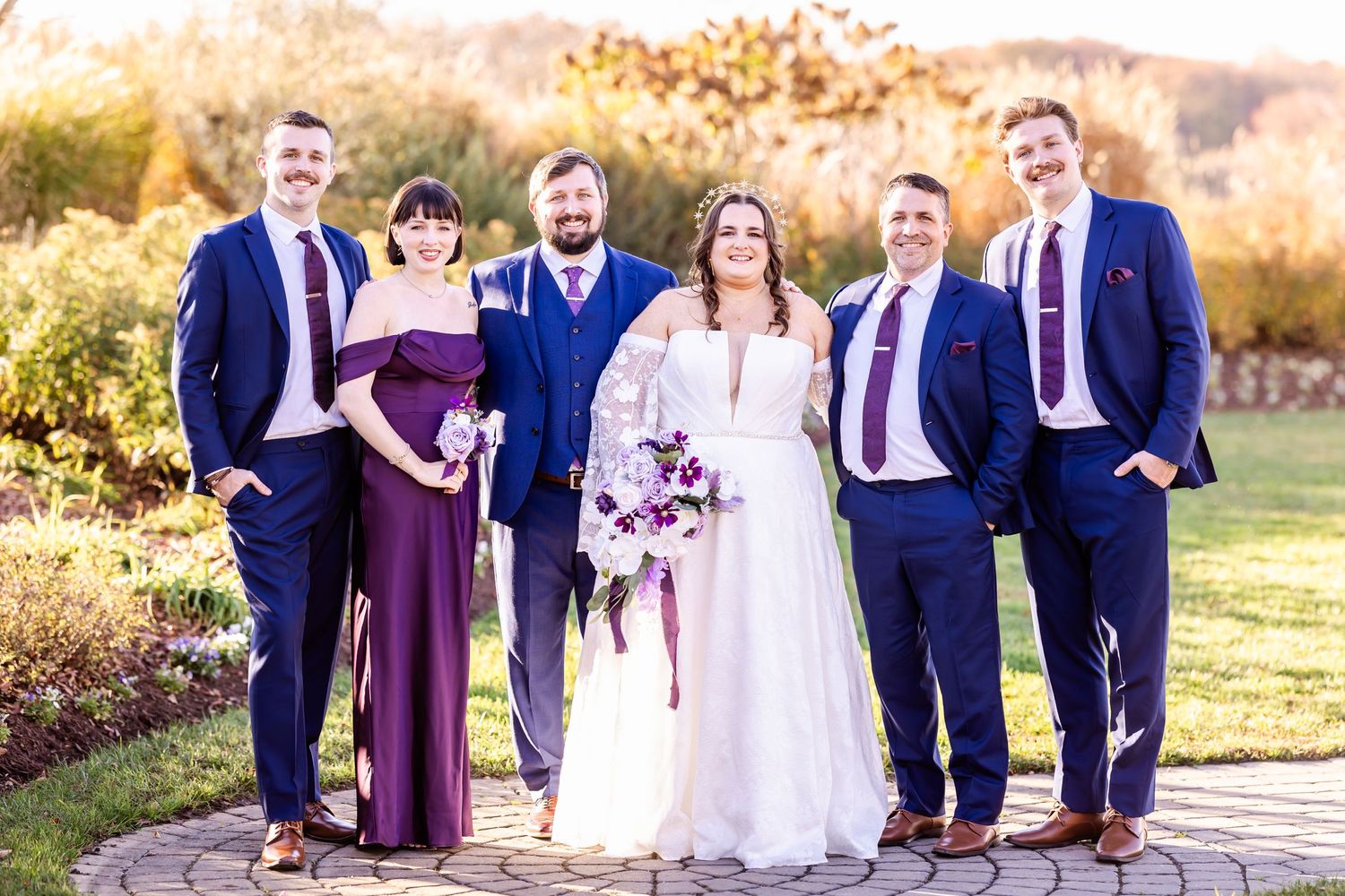 Wedding party in navy suits and burgundy dresses pose with bride in white gown outdoors.