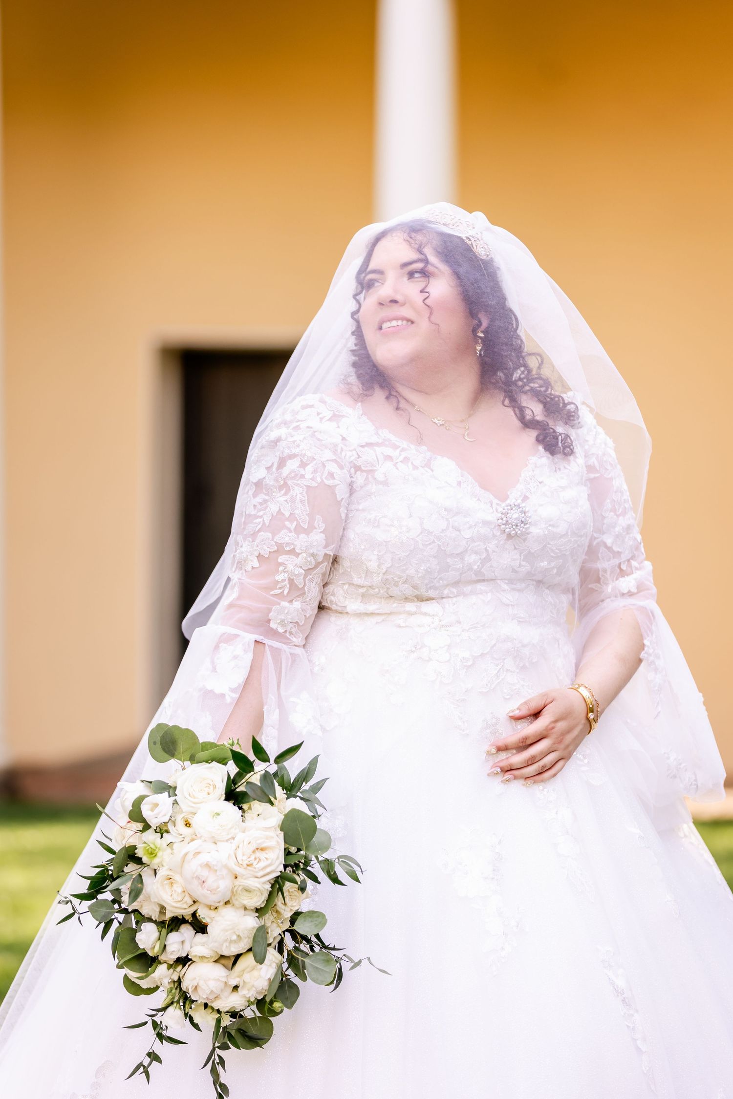 Bride in lace wedding dress holds white floral bouquet against yellow building backdrop.