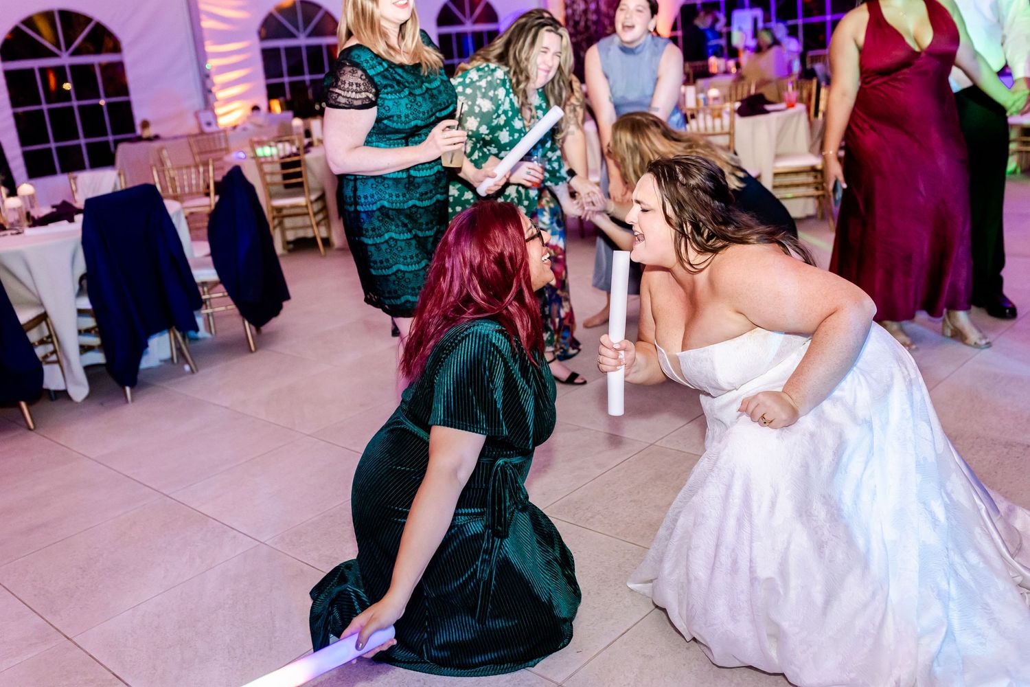 Wedding guests dancing energetically in formal attire under purple-lit tent.