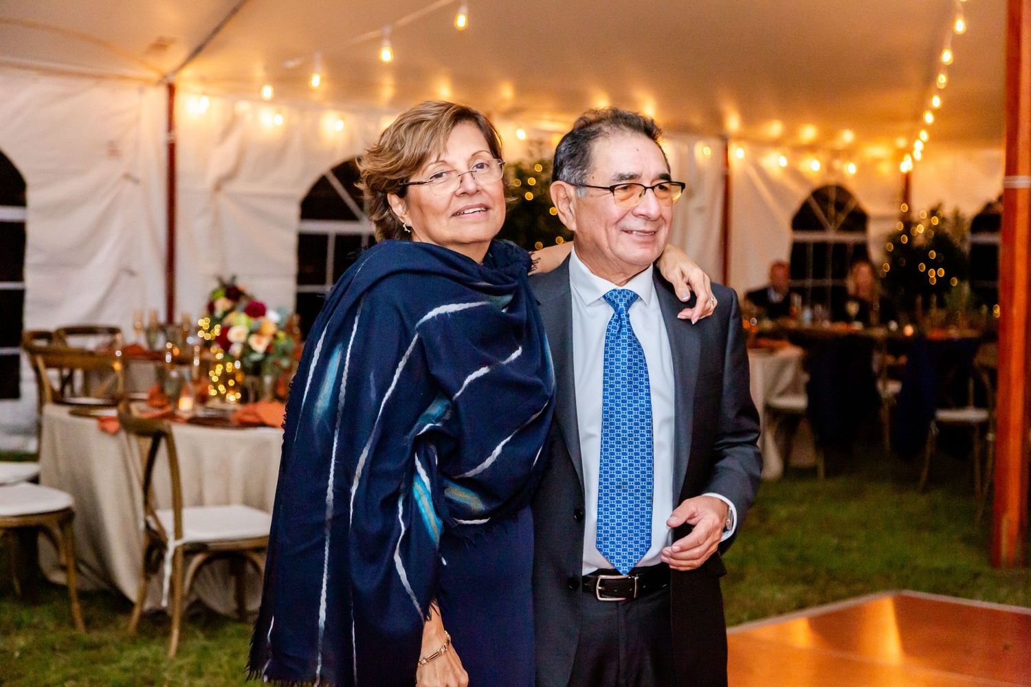 Guests share joyful moments at a wedding reception under warm string lighting in an elegant tent setting.